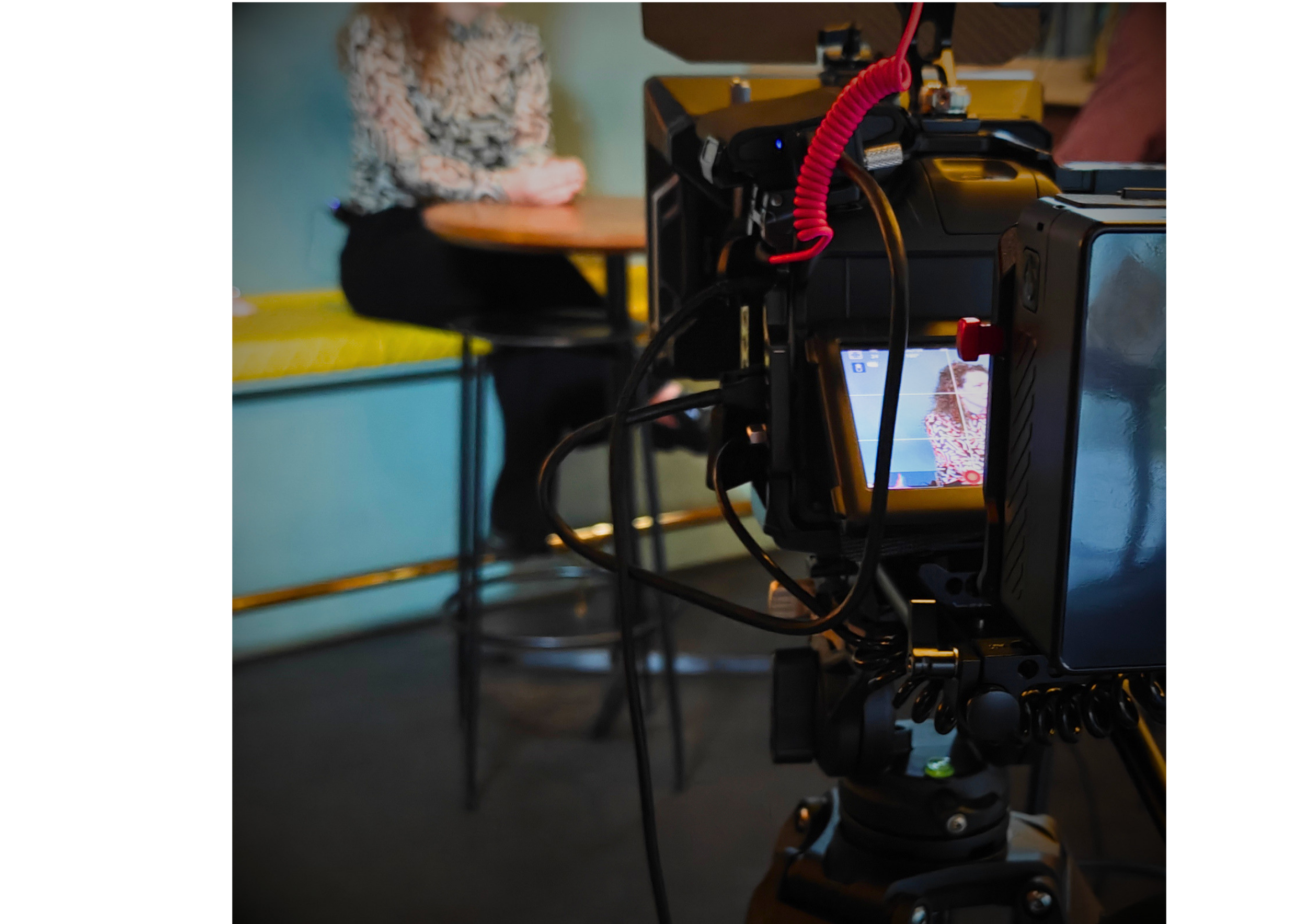 The image shows a camera on a tripod capturing a scene in a room with a person sitting at a table, wearing a floral shirt, with their hands clasped. The person's face is out of focus, and the scene is viewed from behind the camera.