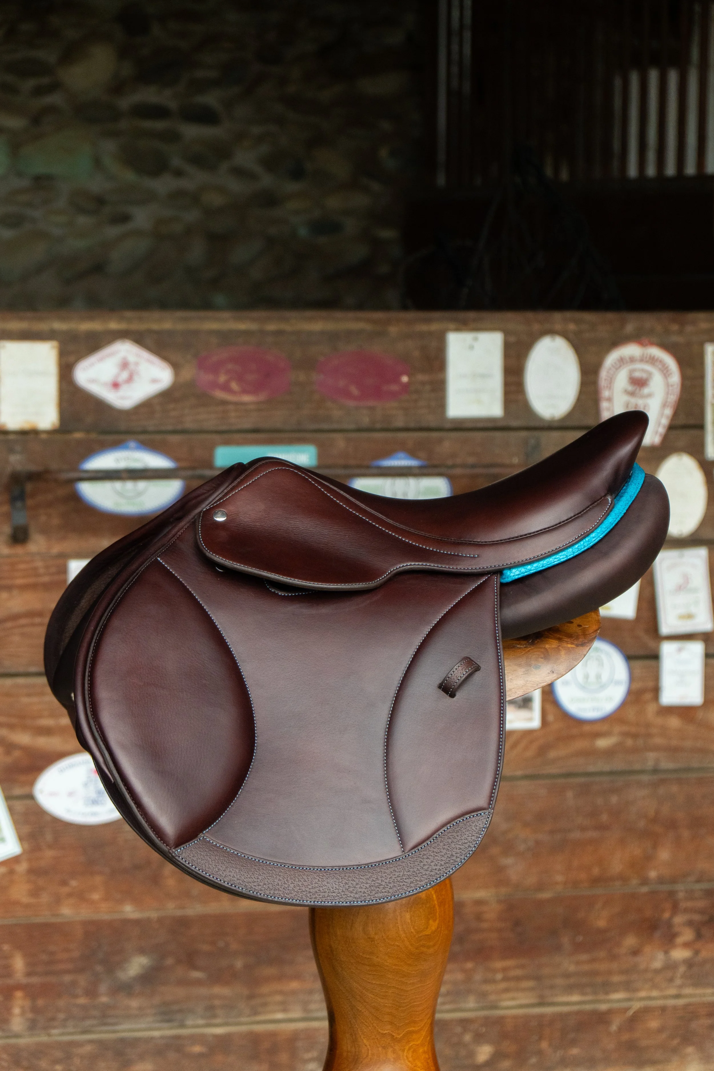 Une selle de cheval en cuir marron posée sur une table en bois avec un fond en pierre et un mur en bois avec plusieurs plaques ou étiquettes.