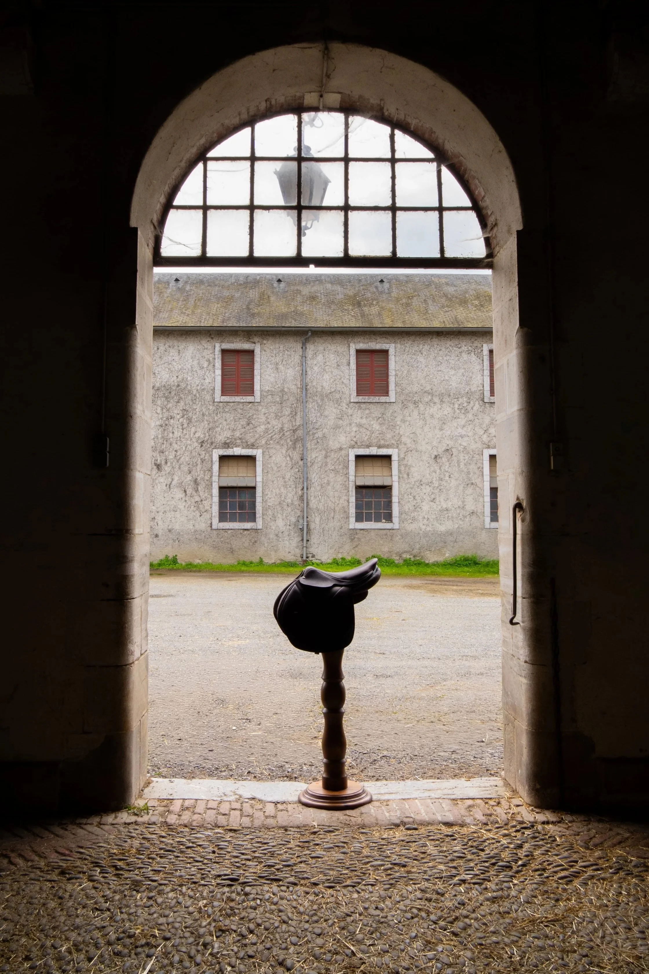 Vue depuis une entrée dans un bâtiment ancien, montrant une cour avec une vieille maison en pierre, fenêtres avec volets rouges, un sol en pavés et une statue de style antique en bois dans le centre.