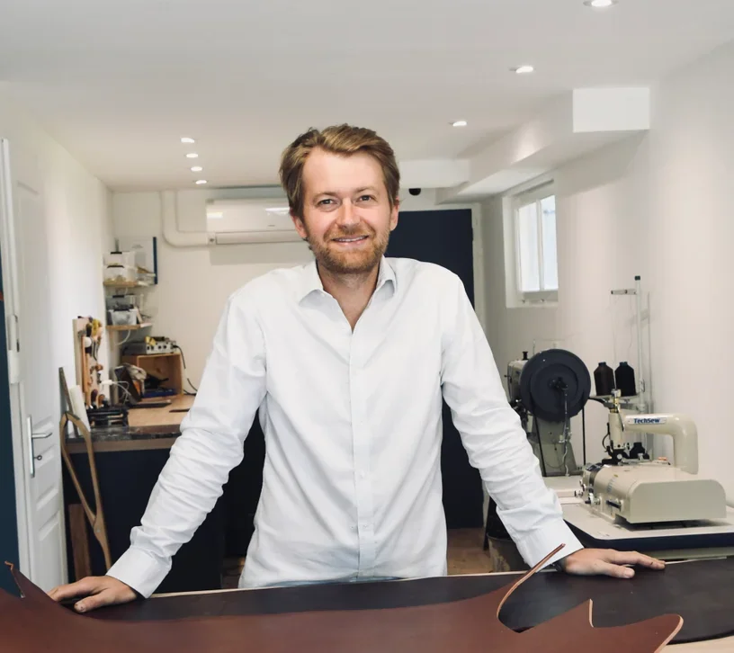 Un homme souriant portant une chemise blanche dans un atelier ou un laboratoire, avec des instruments et machines en arrière-plan.