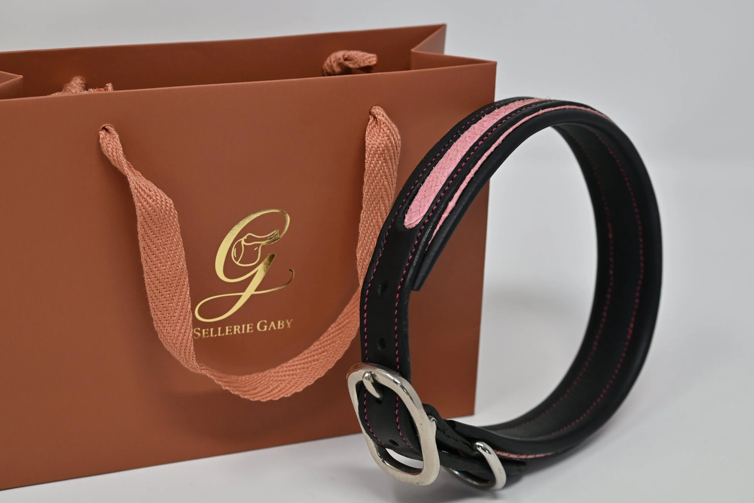 Un sac en papier marron avec des poignées en cordon rose, un logo doré avec un dessin d'une main, et une laisse noire avec des détails roses attachée à un fermoir en métal argenté.