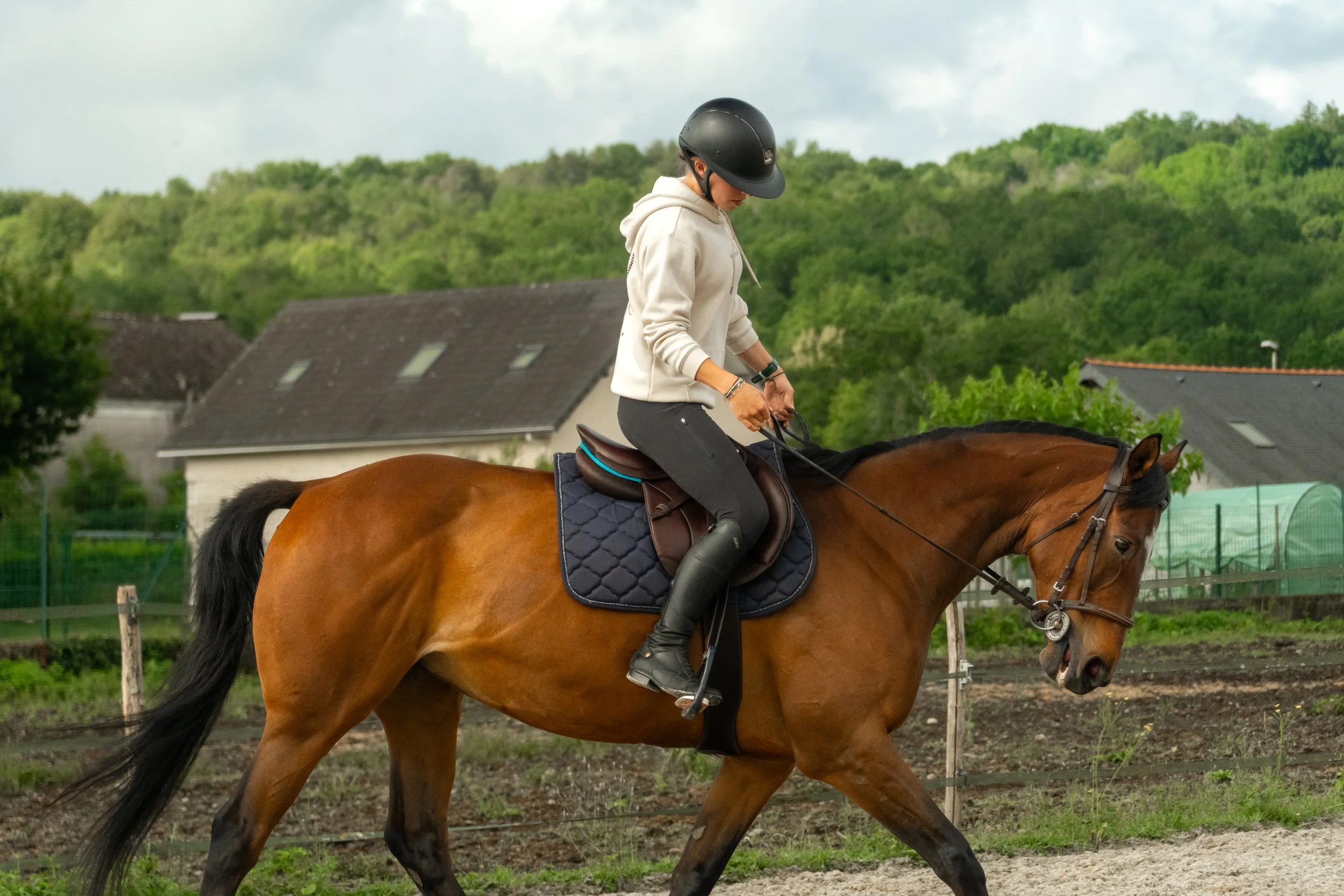 Femme à cheval portant un casque noir, sweat à capuche beige, dans un enclos avec des bâtiments et une végétation verte en arrière-plan.