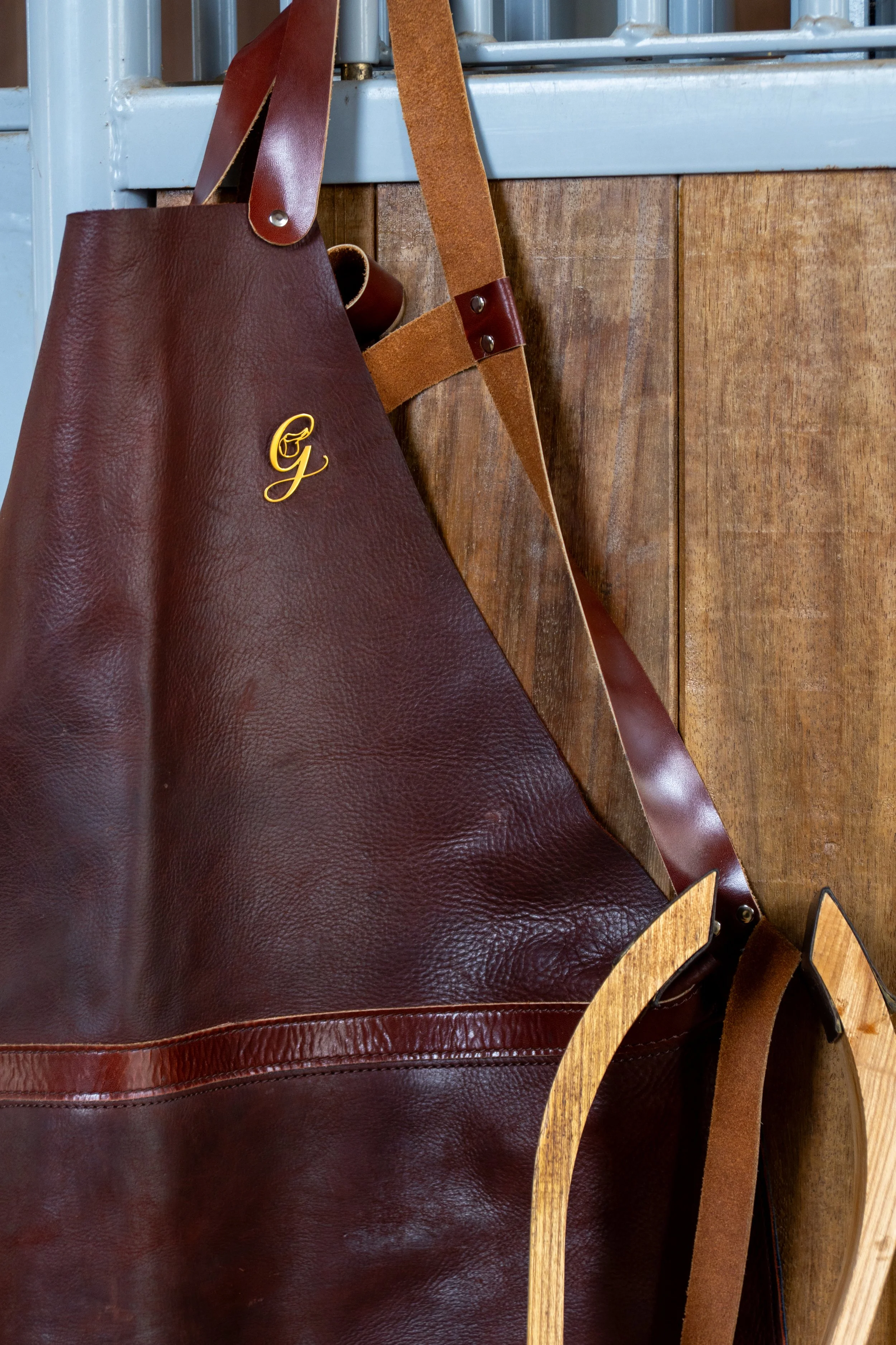 Une grande sacoche en cuir marron avec des détails en cuir plus clair et une broderie dorée. Elle est suspendue à une balustrade en métal, en face d'un panneau en bois.