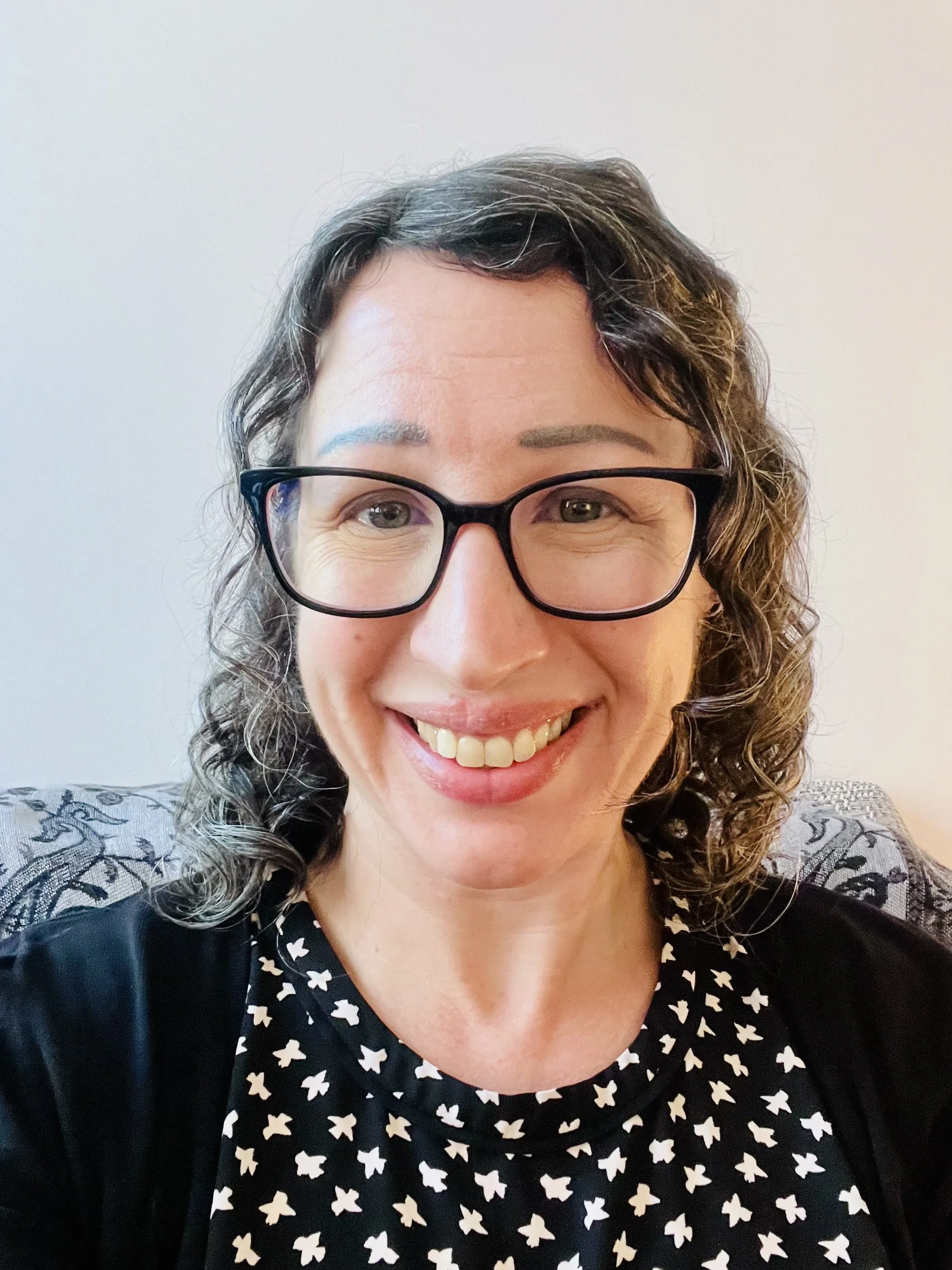 Close-up photo of a smiling woman with curly brown hair, wearing black-rimmed glasses and a black dress with white star patterns.
