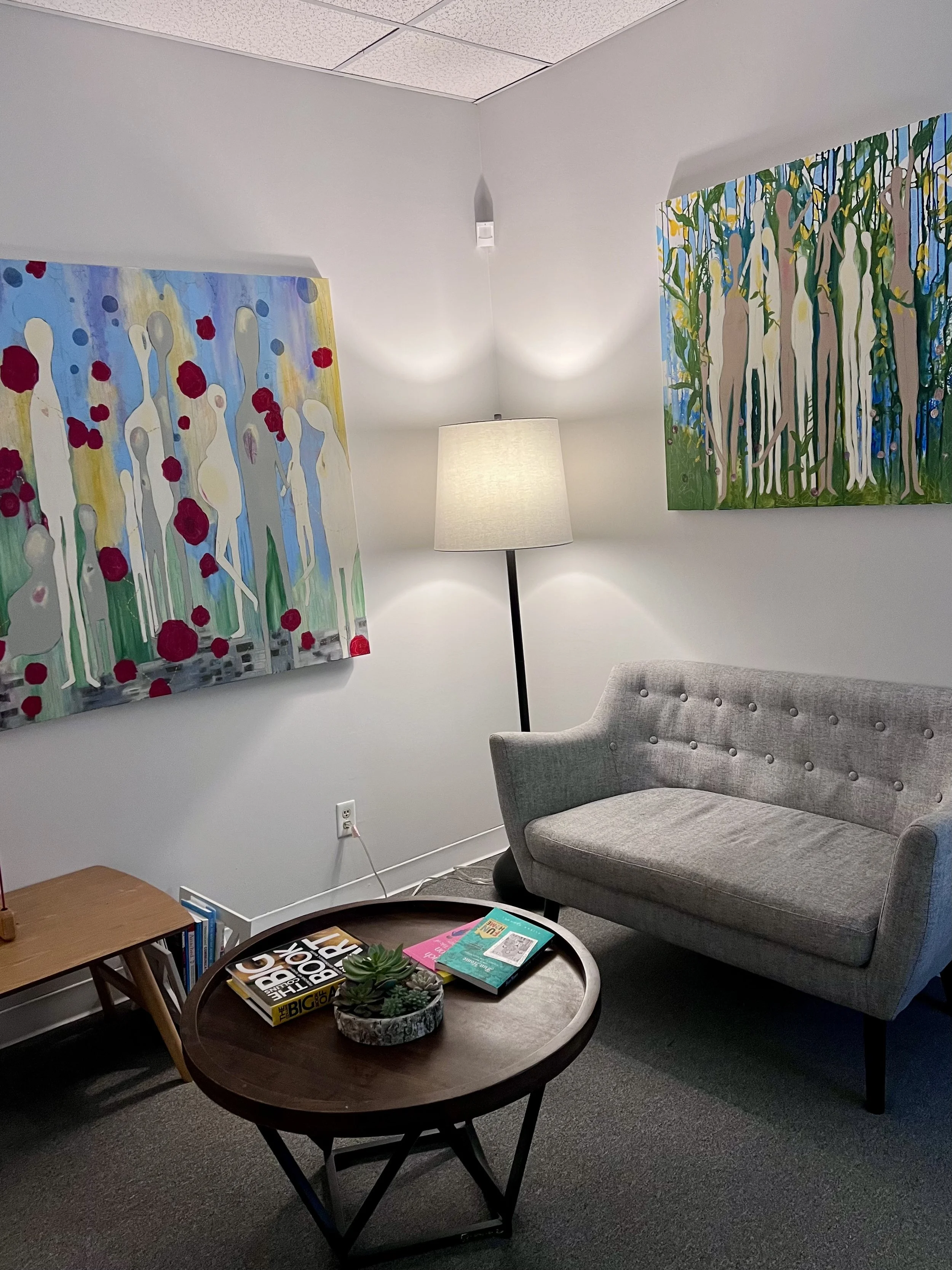 A corner of a cozy living room with a tripod table holding magazines and succulents, a gray armchair, a wooden side table with books, two colorful abstract paintings on the white walls, a floor lamp, and a textured carpet.