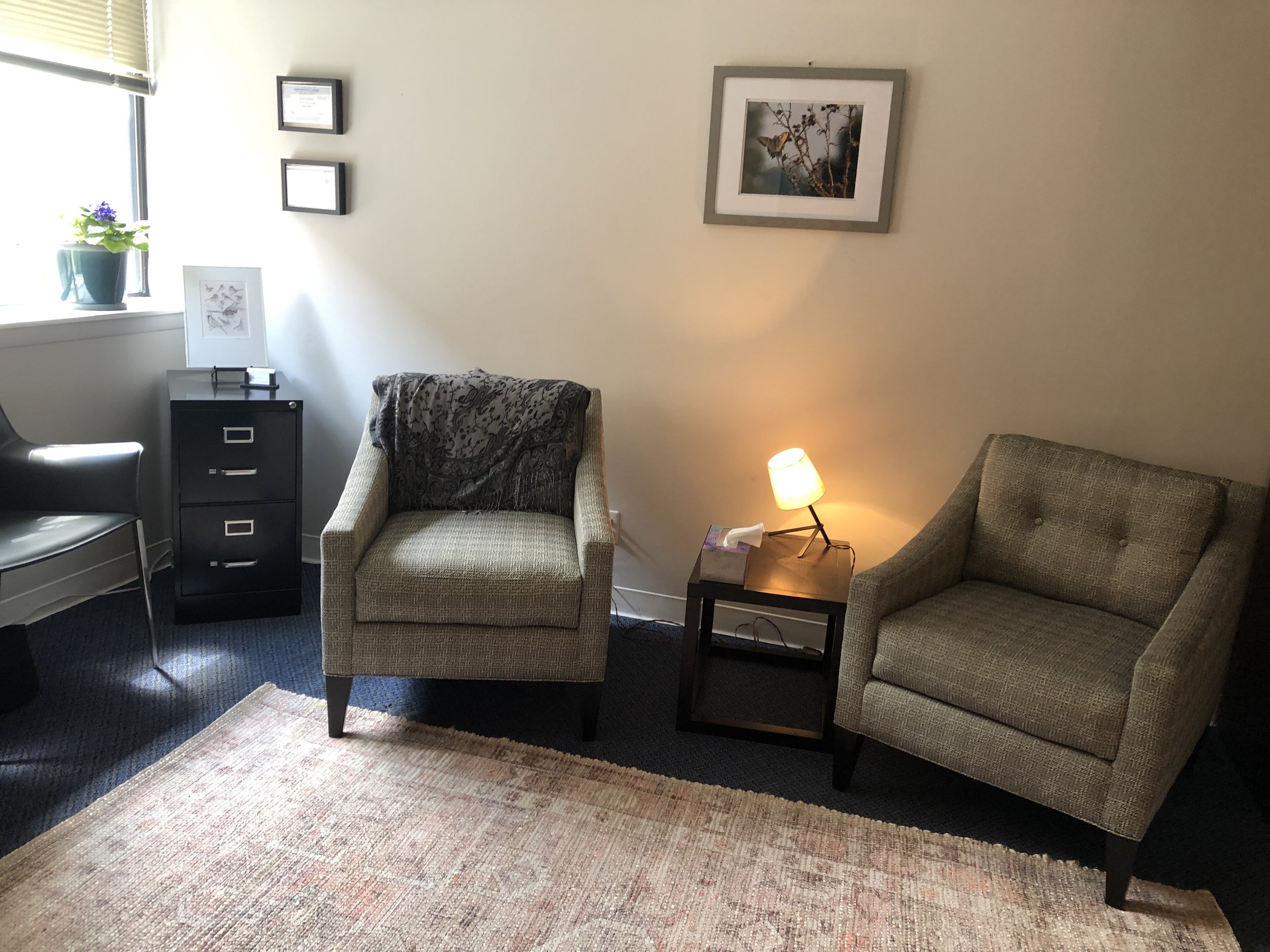 A therapy office with two armchairs, a black side table with a lamp, a window with blinds, and framed pictures on the wall. There is a small potted plant on the window sill.