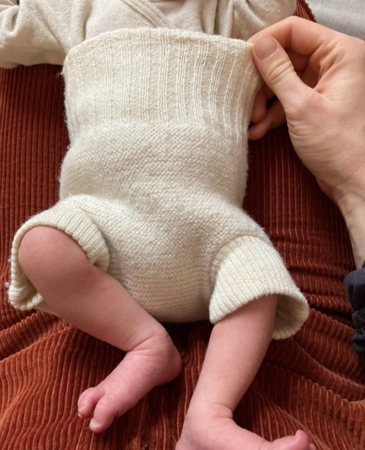 Nahaufnahme eines Babys in einem hellen Pullover, gehalten von Erwachsenen, die auf einem braunen, gestrickten Stoff sitzen.