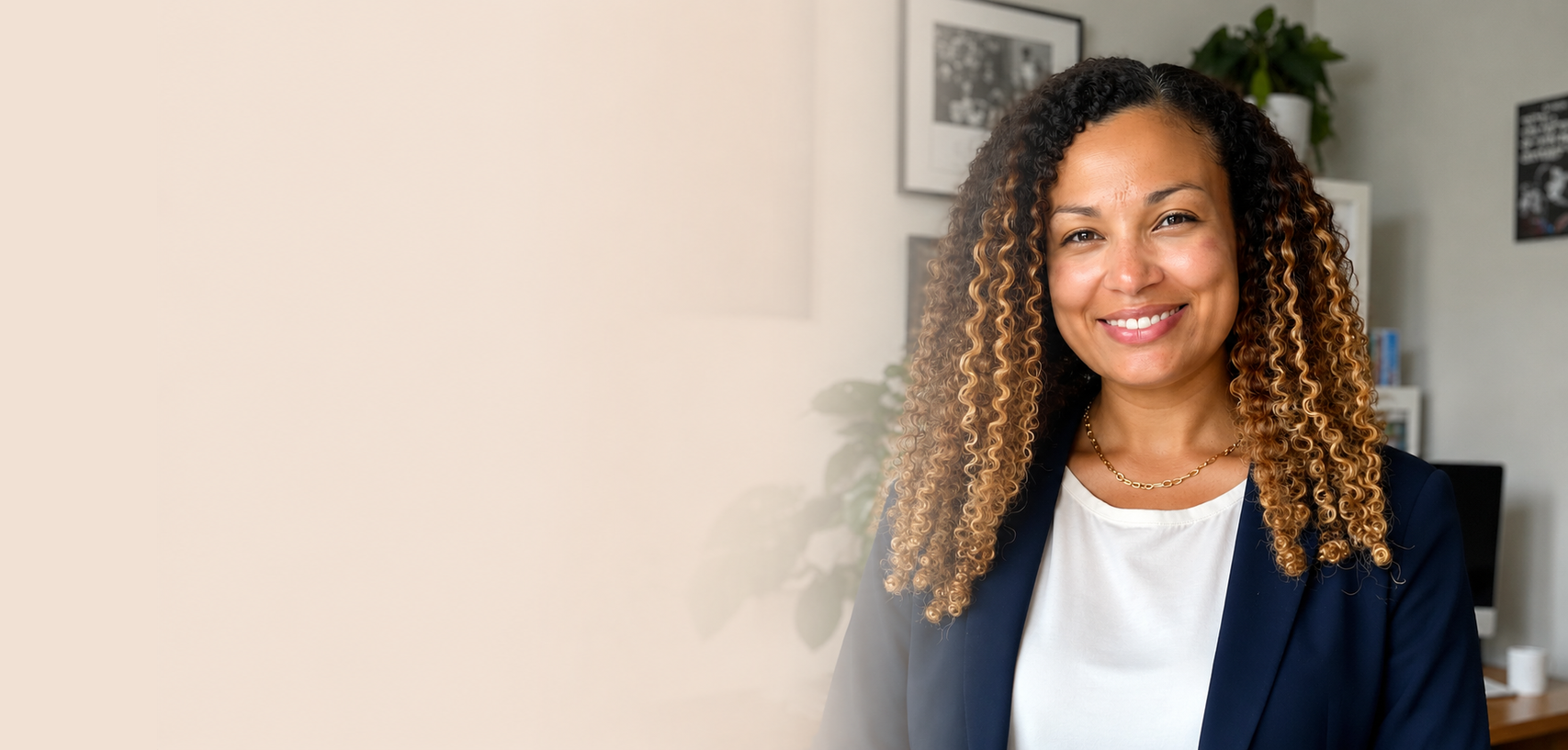 Smiling woman with curly hair wearing a navy blazer and white shirt standing in an office with framed pictures and a computer in the background.