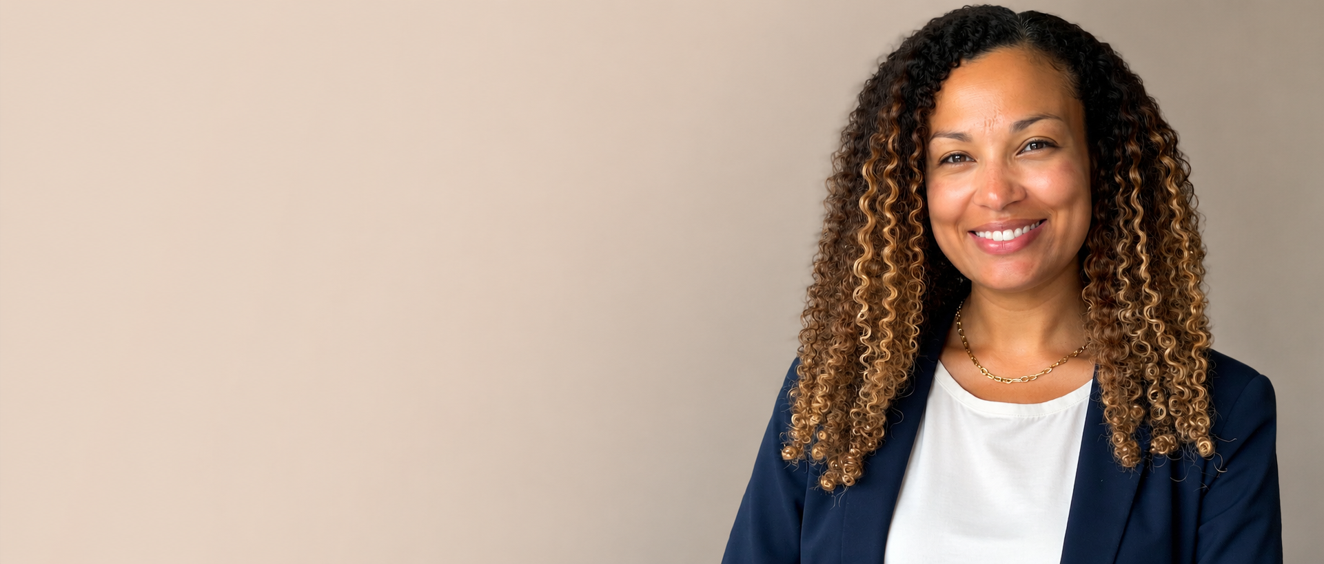 A smiling woman with curly hair wearing a navy blazer and white top, standing against a neutral background.