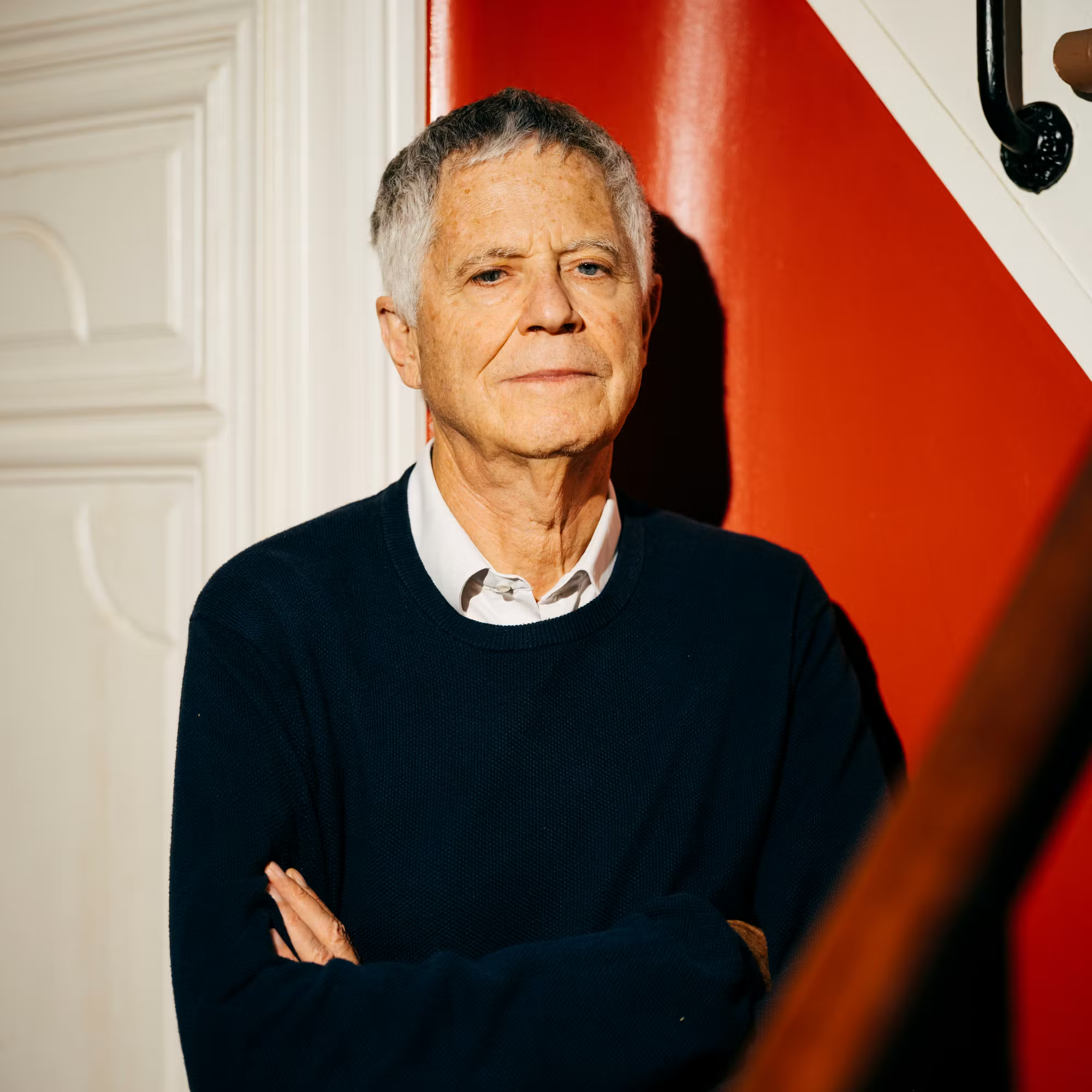 Portrait d'un homme âgé avec des cheveux gris, portant un pull noir et une chemise blanche, debout contre un mur rouge avec un décor blanc, bras croisés.
