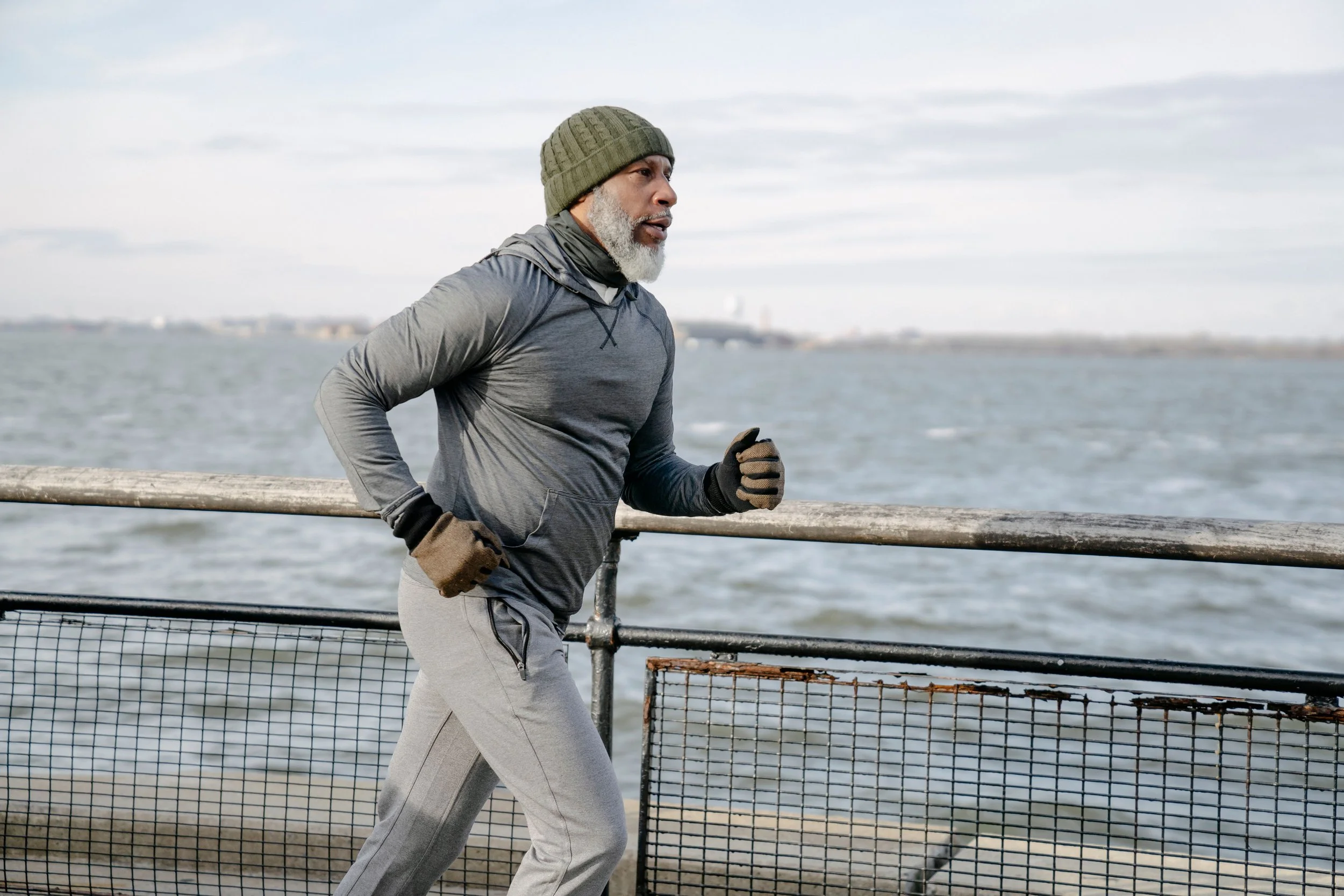An older man with a gray beard running outdoors near a body of water, wearing a gray jacket, beige running pants, gloves, and a knit beanie.