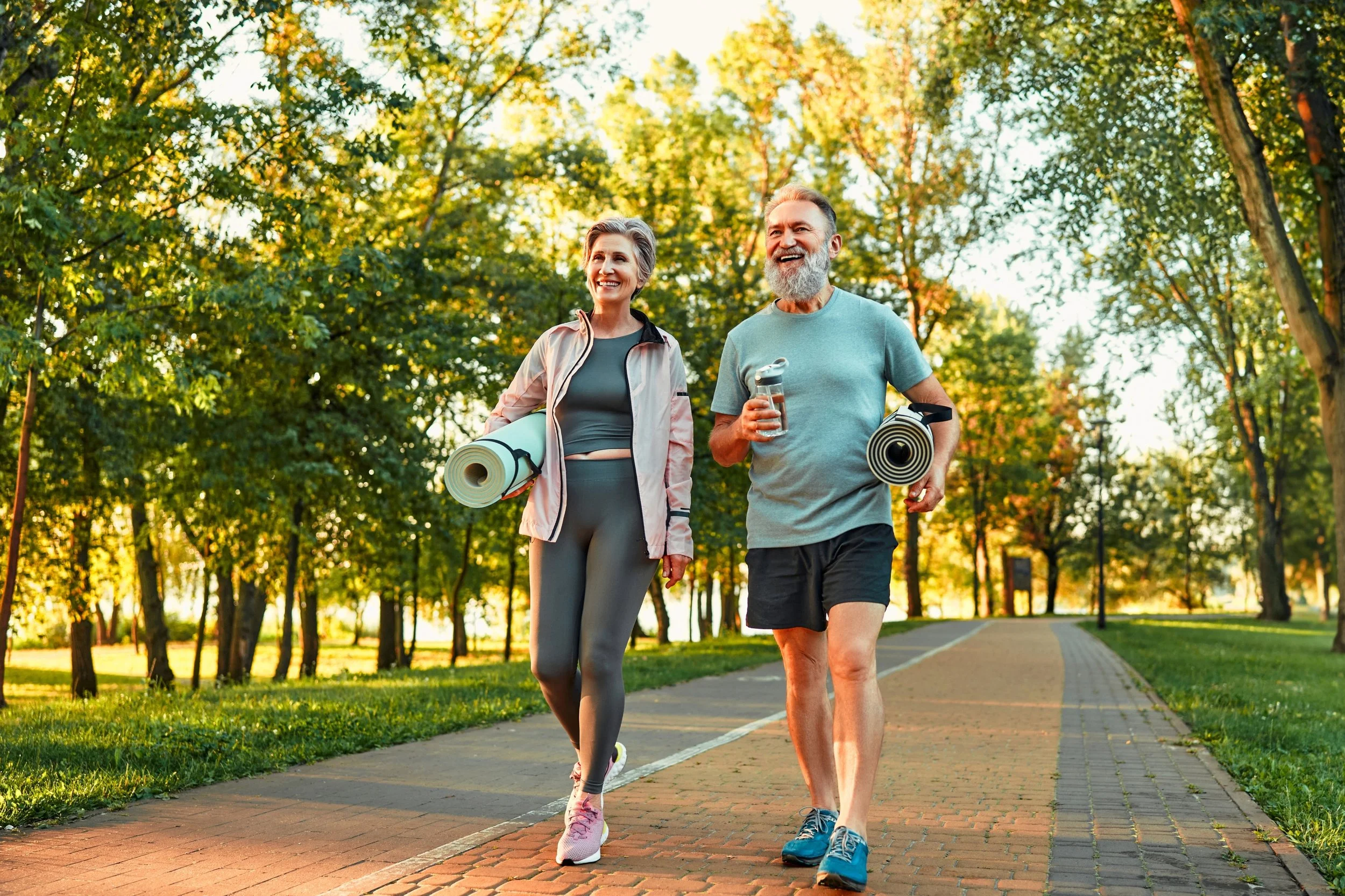 An elderly man and woman walking in a park, smiling, with trees in the background. The woman carries a yoga mat, and the man holds a water bottle and a yoga mat. They are dressed in athletic clothing.