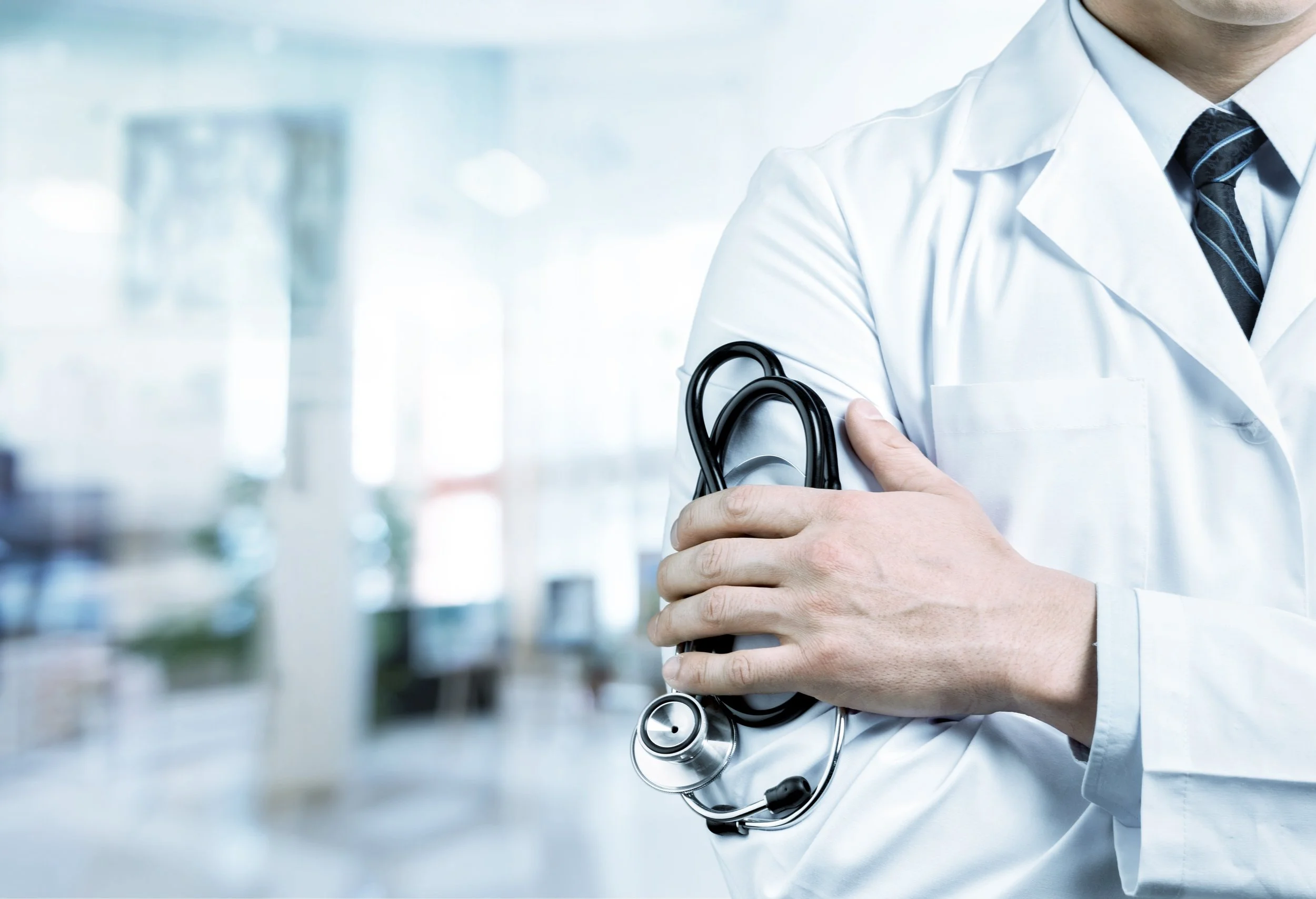 Close-up of a medical doctor wearing a white coat and stethoscope with arms crossed, in a hospital or clinic setting.