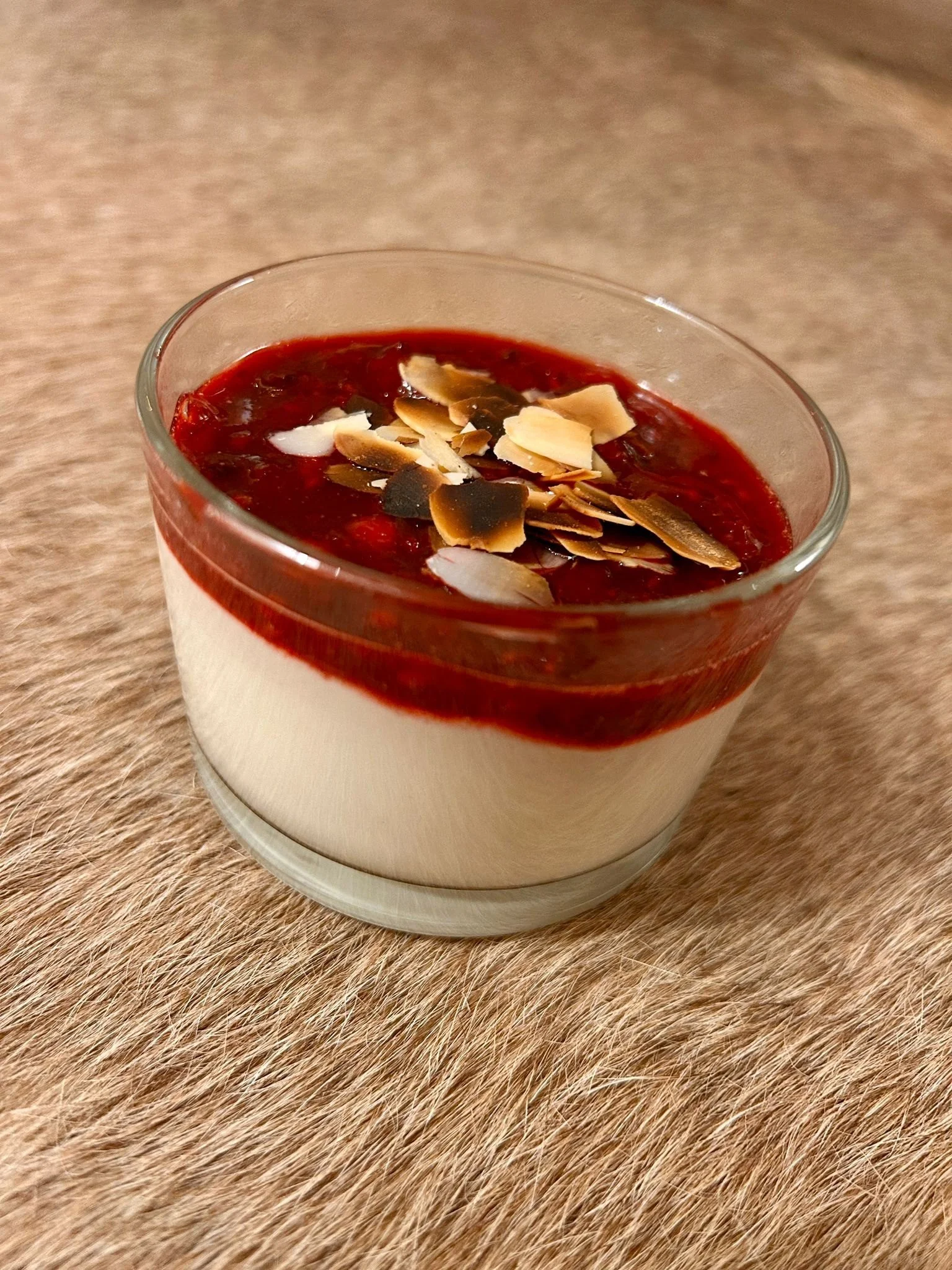 A glass of layered dessert with white base, red fruit topping, and toasted sliced almonds on top, placed on a textured brown surface.