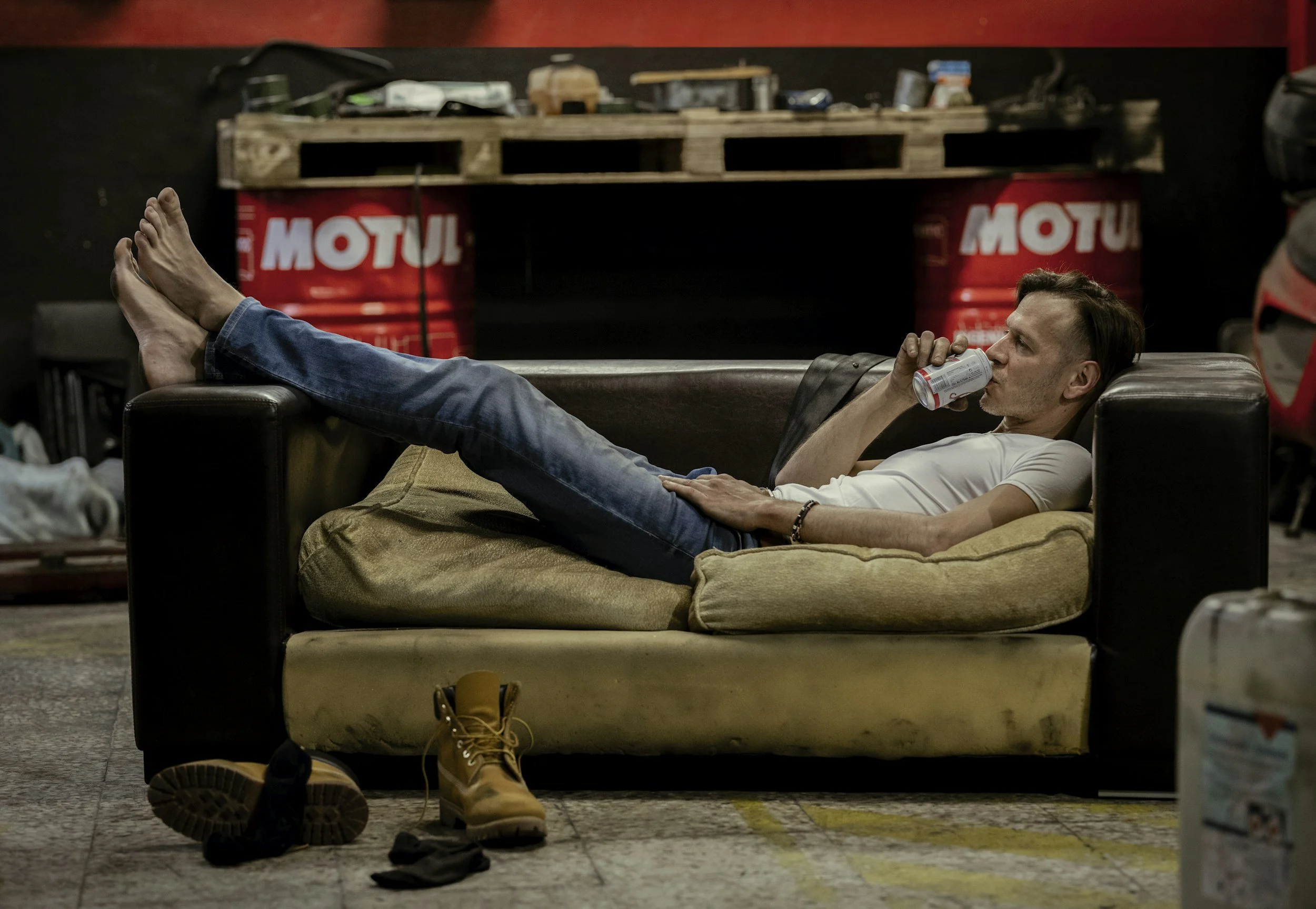 A man relaxing on a couch, drinking from a can, with his feet resting on the armrest. The background features a workbench with various tools, two large red barrels labeled 'MOTUL', and a pair of boots on the floor.