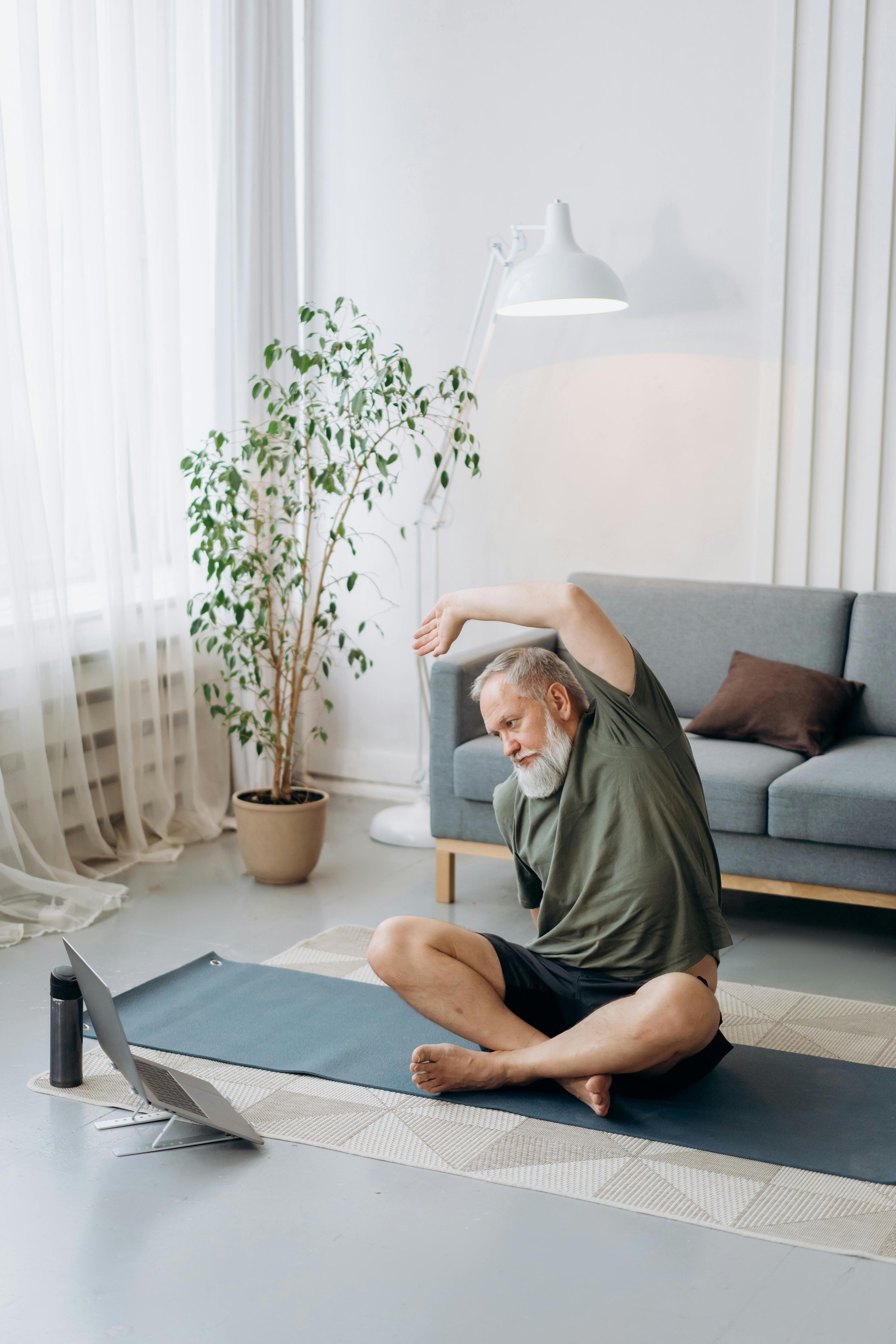 An elderly man with gray hair and a beard practicing yoga or stretching in a bright living room. He is sitting cross-legged on a yoga mat with one arm extended overhead and the other touching his foot. There is a tablet propped on a stand, a water bottle, and a laptop nearby. The room has a large window with sheer curtains, a potted plant, a gray sofa, and a white floor lamp.