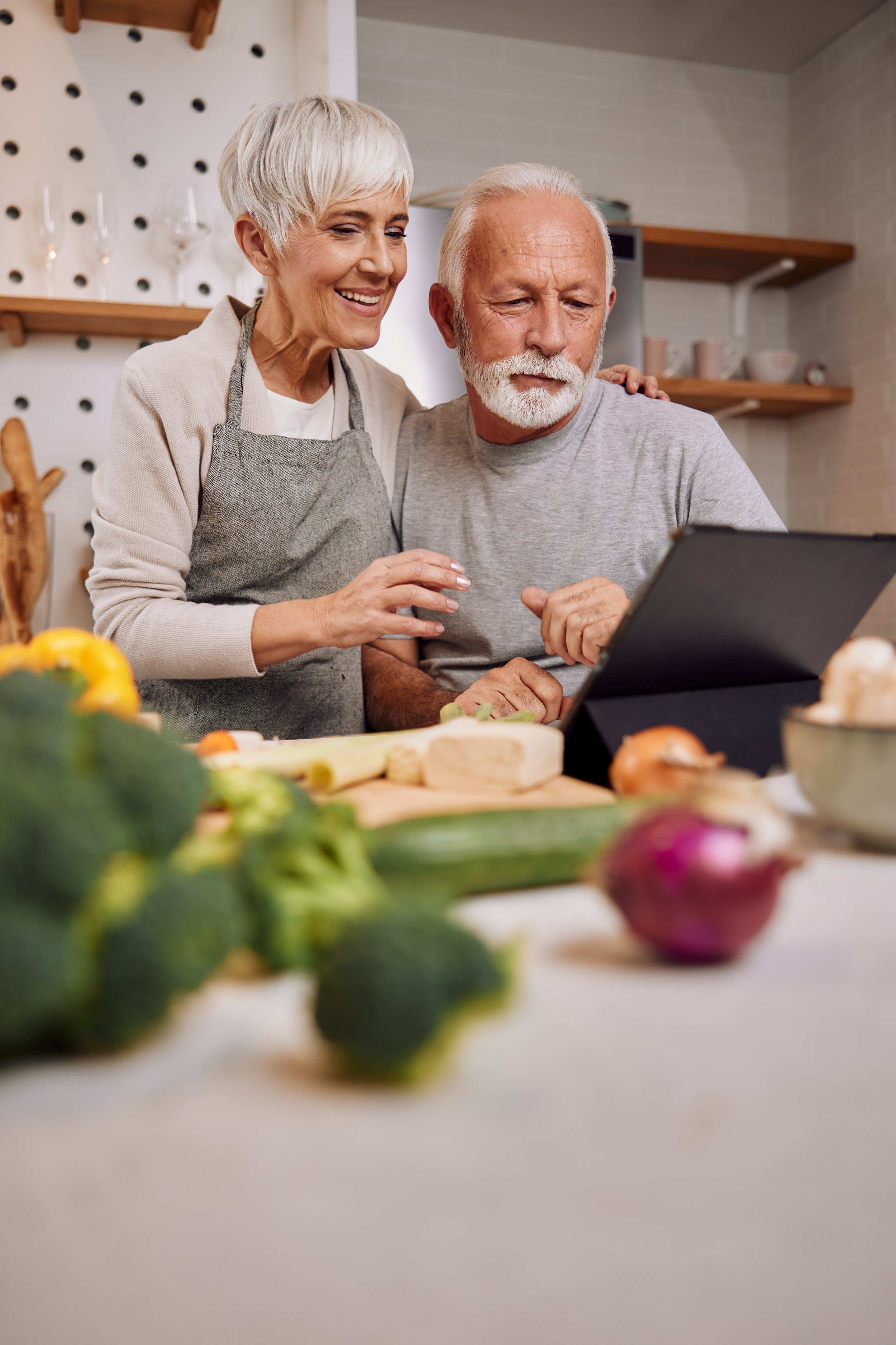 A happy elderly couple shopping for vegetables in a kitchen, with various vegetables on the table in the foreground.