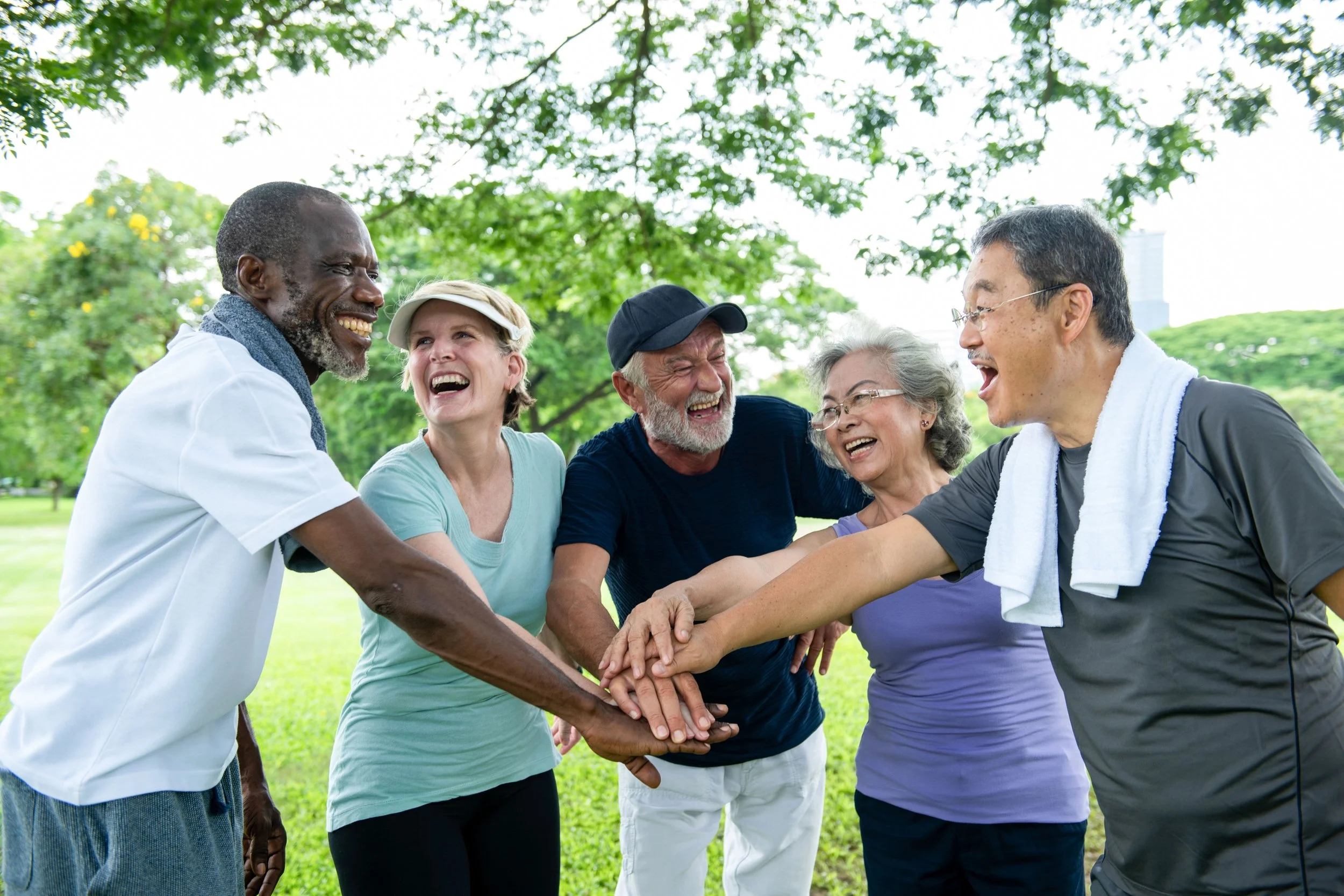 A diverse group of six people, including men and women of varying ages, are standing outdoors under a large green tree, smiling and with their hands stacked in the center, engaging in a group cheer or team-building activity in a park.