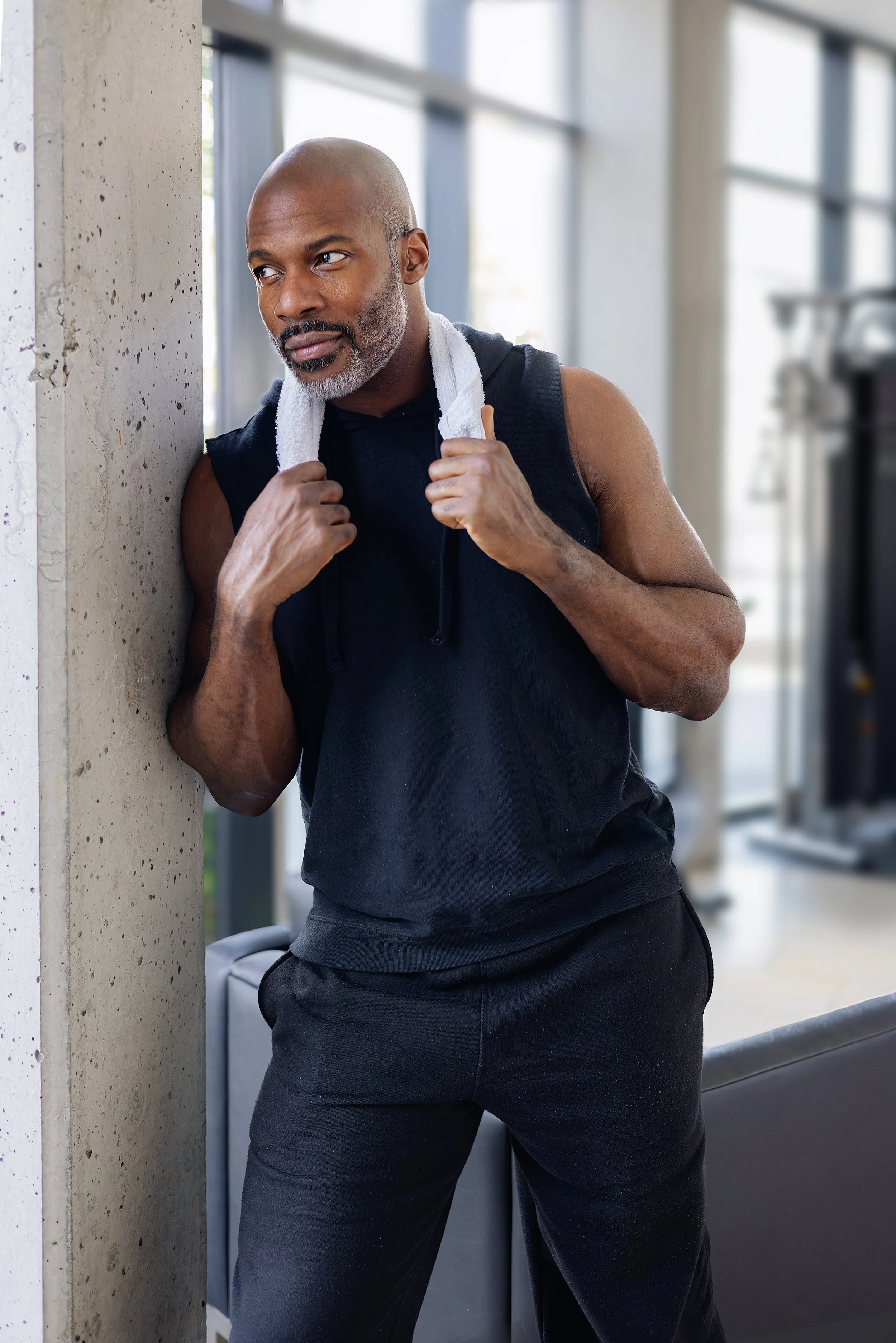 A muscular bald man with a gray beard stands in a gym, leaning against a concrete wall, holding a white towel around his neck, dressed in a black sleeveless shirt and black pants.