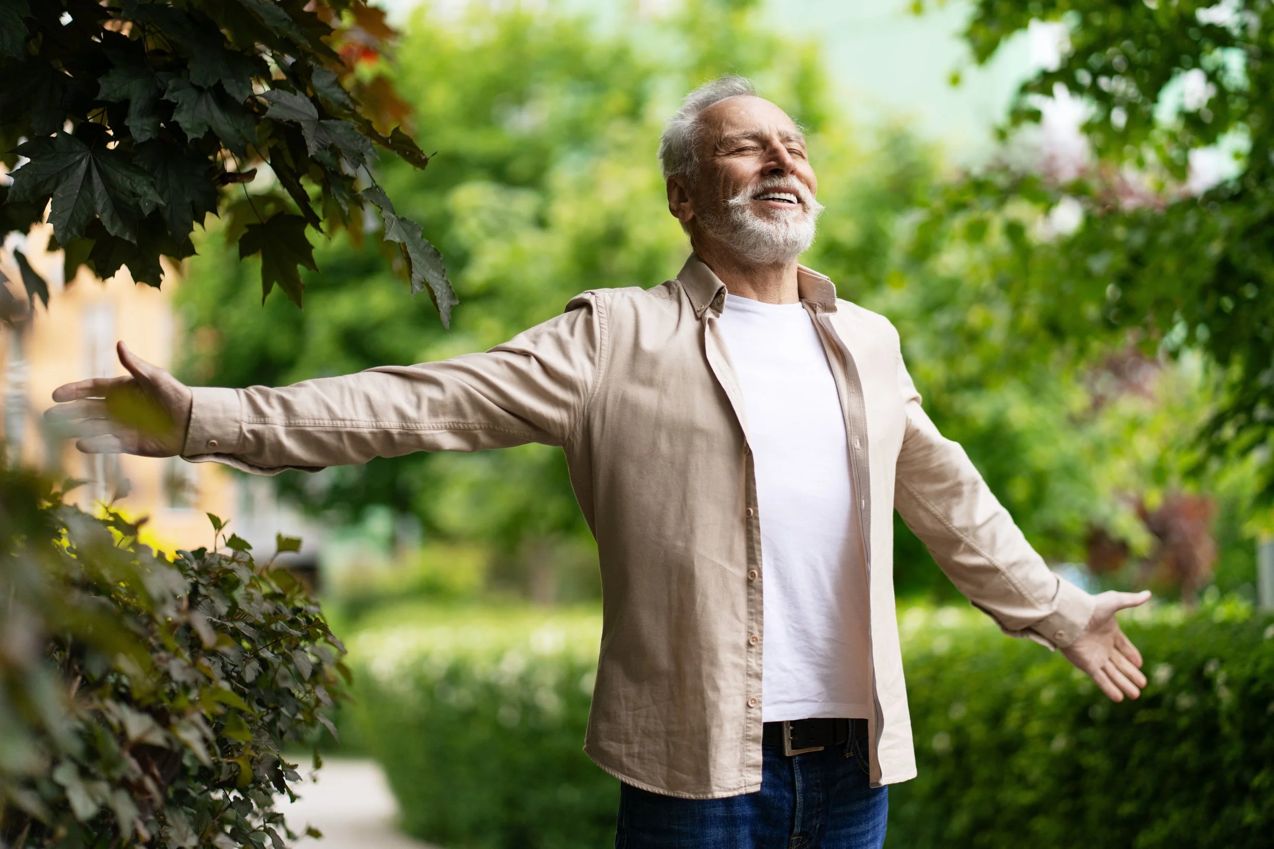 An older man with gray hair and a beard smiling with arms outstretched in a park with green trees and bushes.