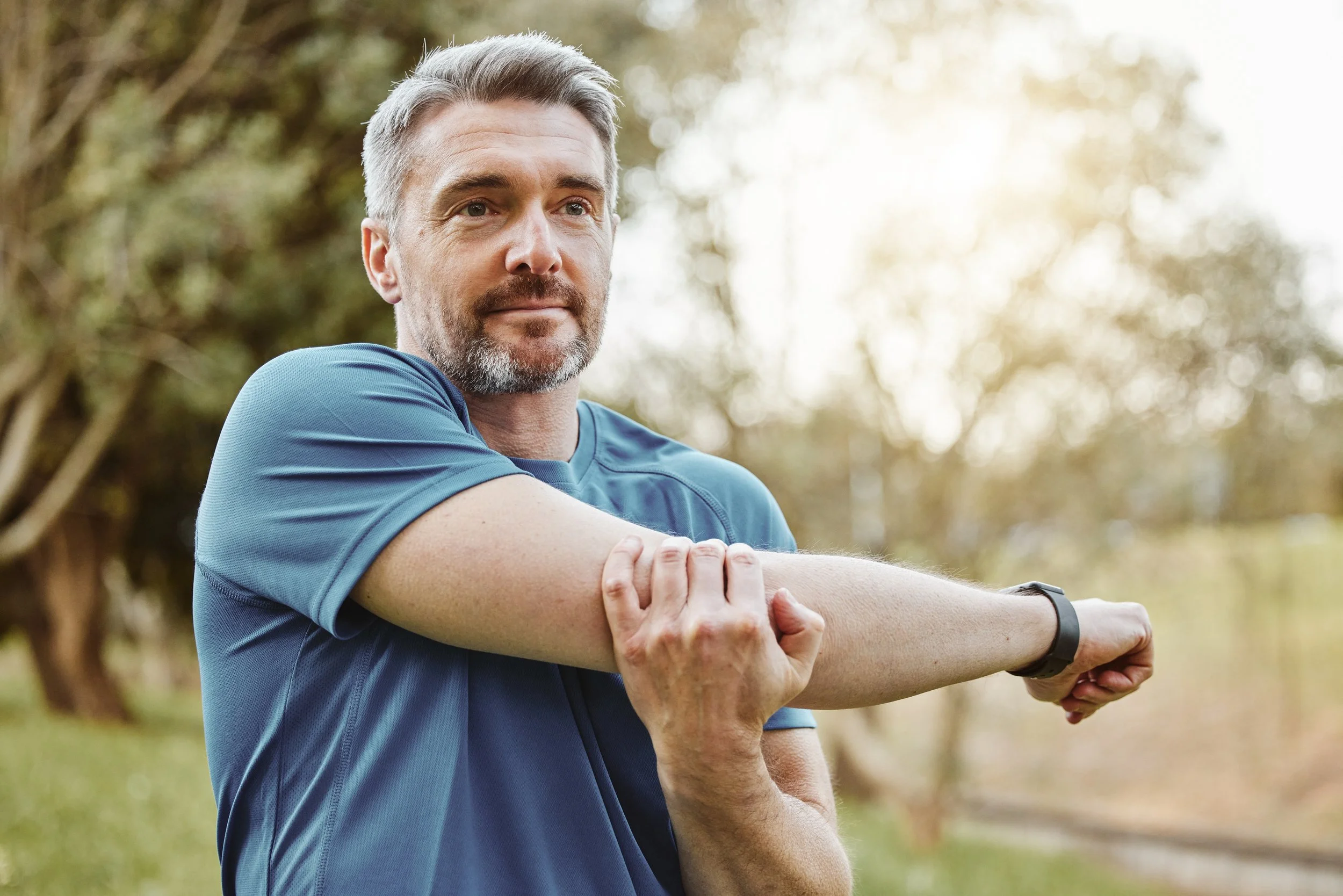 A middle-aged man with gray hair and a beard stretches his arm outdoors in a park, with trees and sunlight in the background.