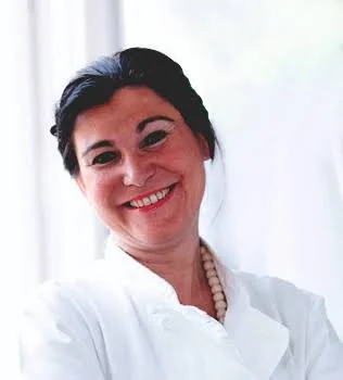 Portrait of a woman smiling, wearing a white top and a pearl necklace, with dark hair pulled back, standing in front of a light background.