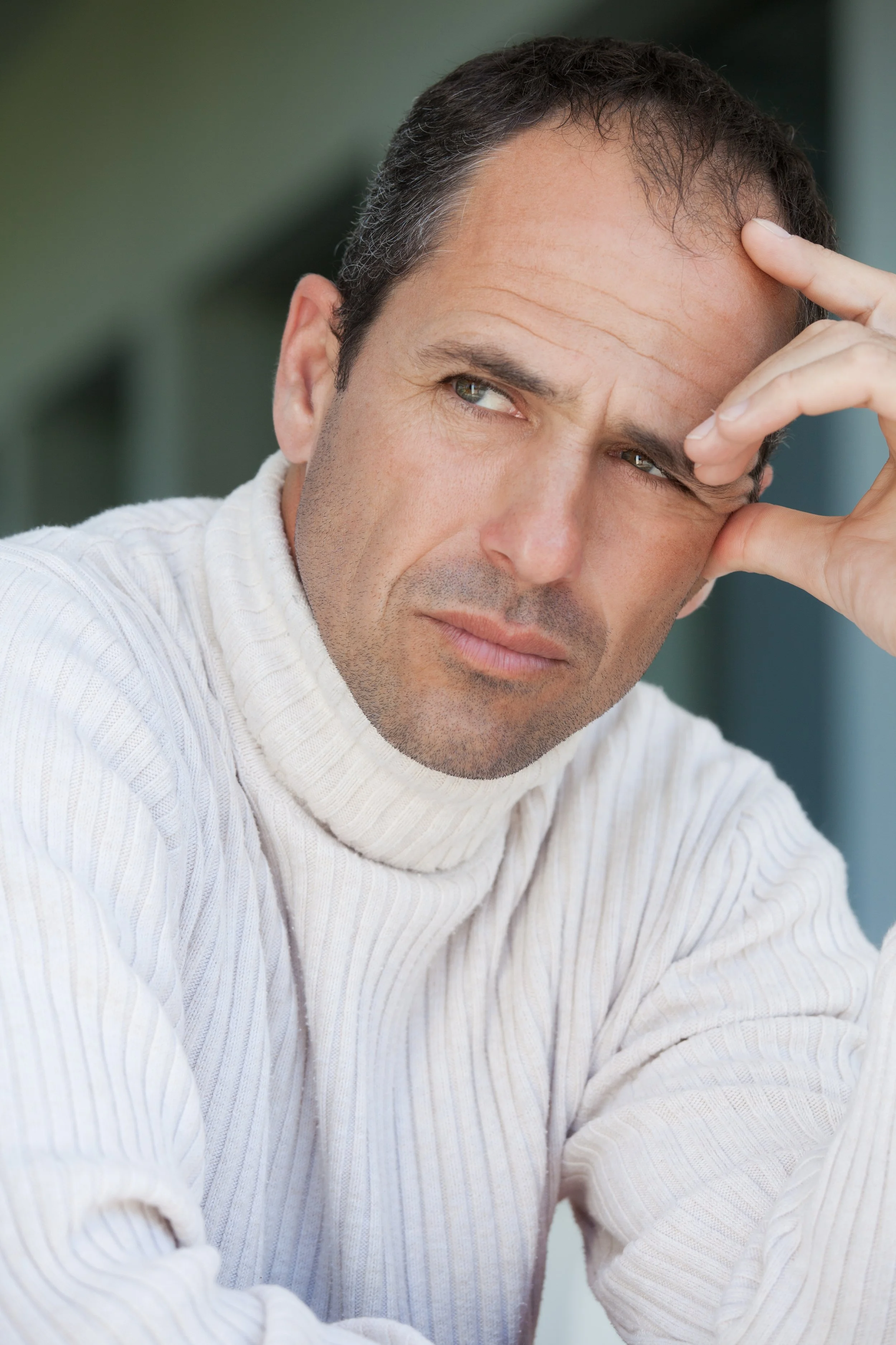 A man with dark hair and light skin, wearing a white turtleneck sweater, resting his head on his hand and looking to the side with a thoughtful expression.