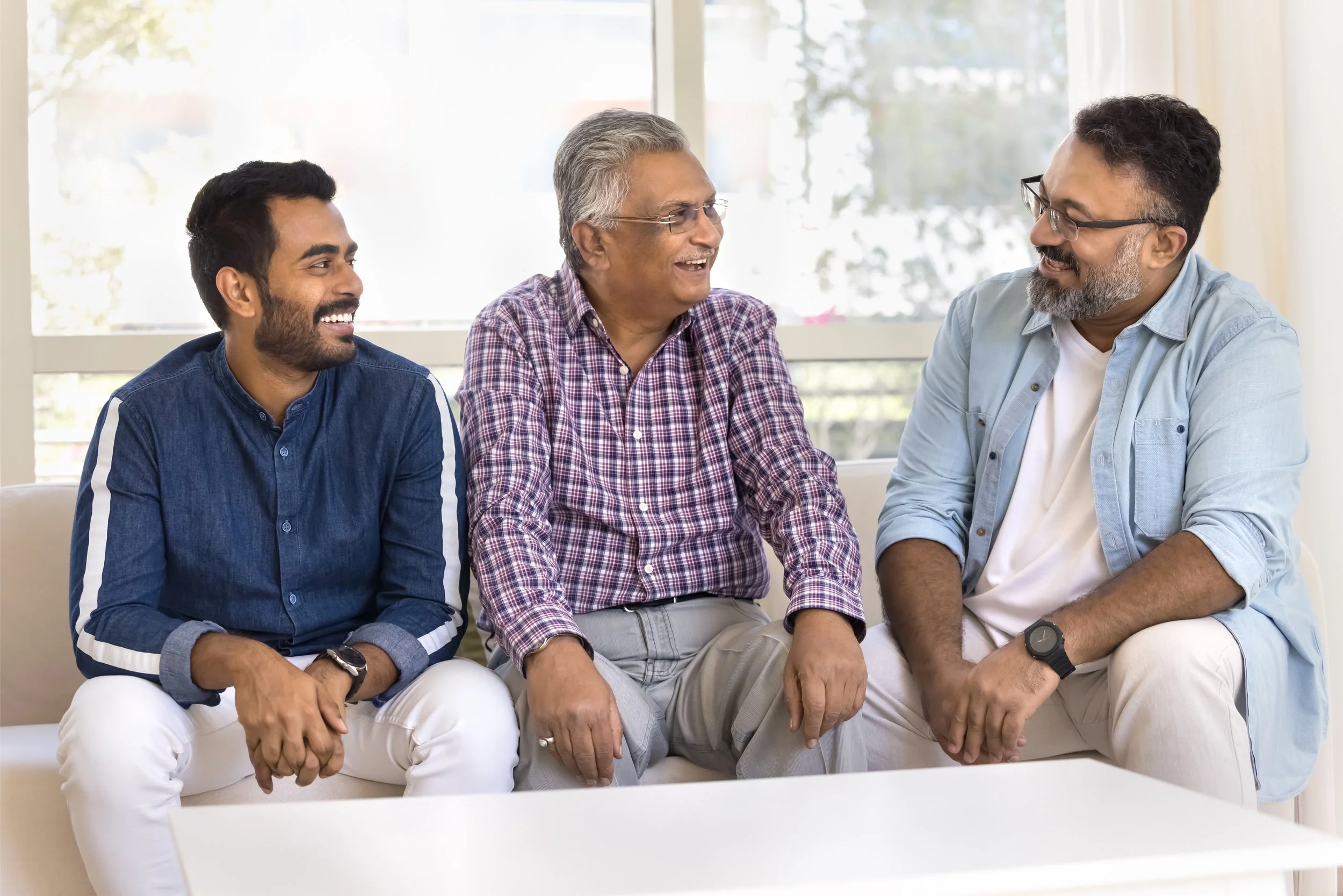 Three men sitting on a white sofa, engaged in conversation and smiling, in front of large windows with sunlight.