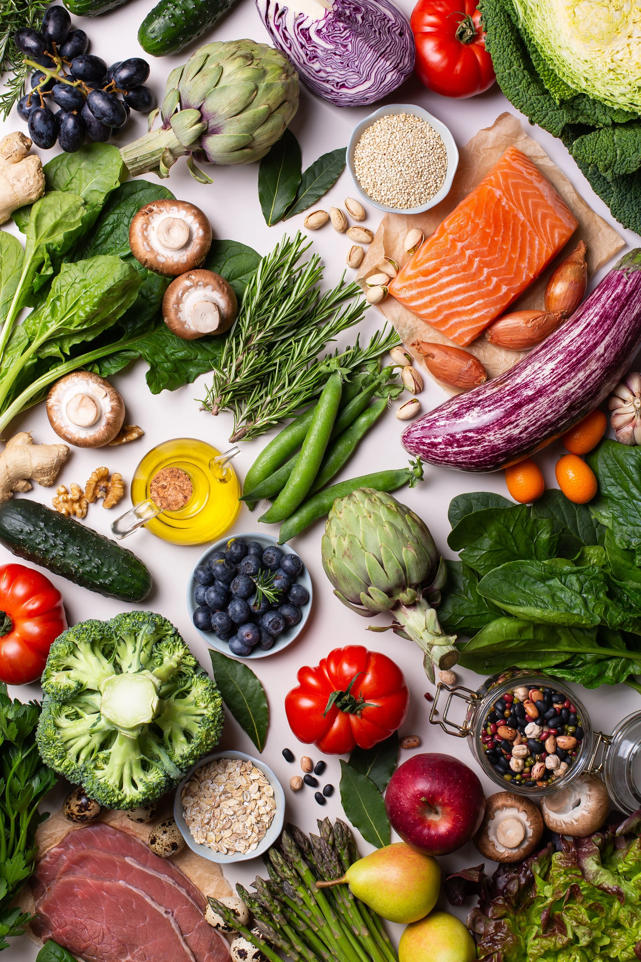 Assortment of fresh vegetables, fruits, fish, nuts, and grains on a white surface.