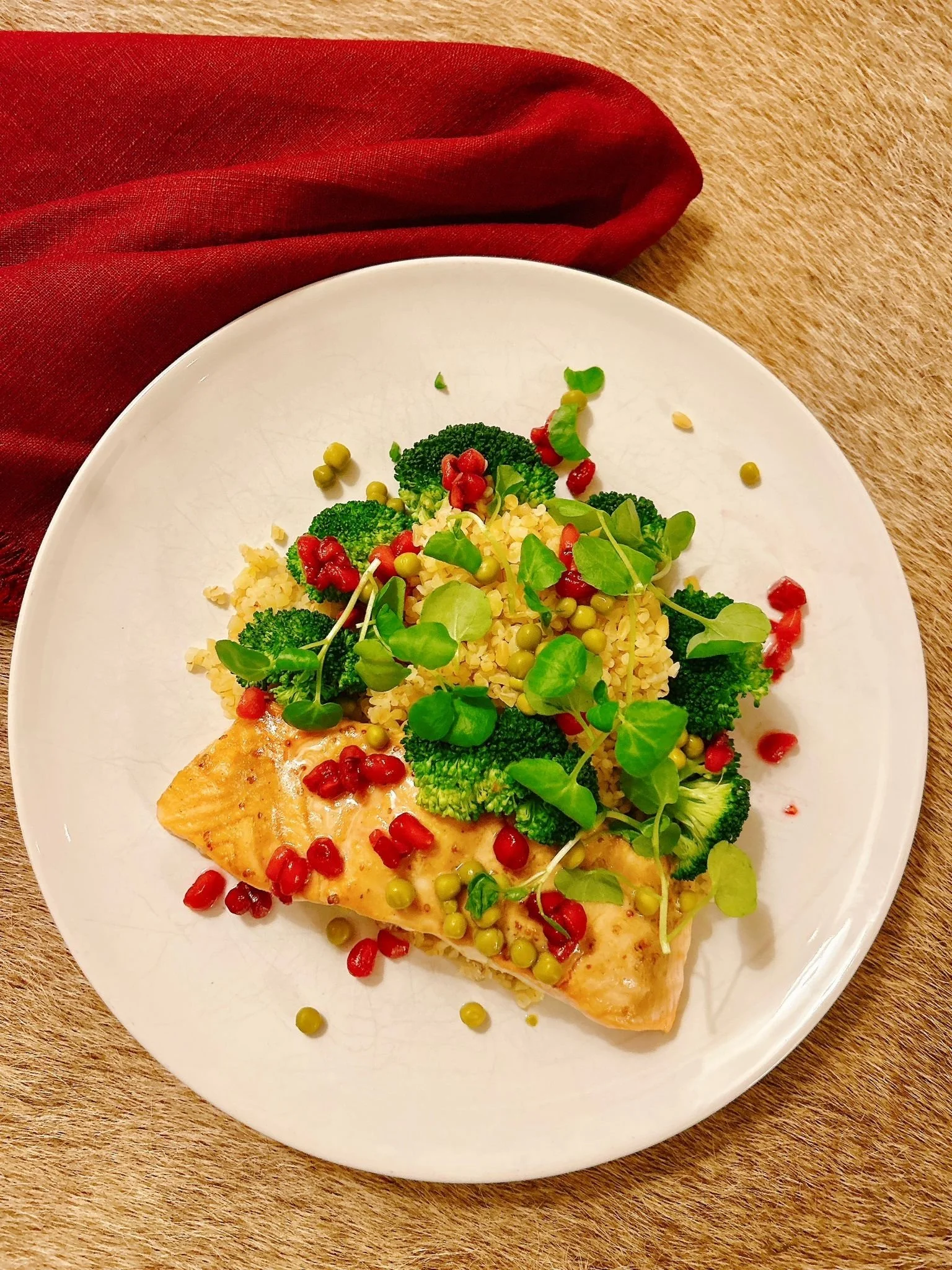 A white plate with baked fish topped with green peas, microgreens, broccoli, and pomegranate seeds, on a wooden table with a red napkin