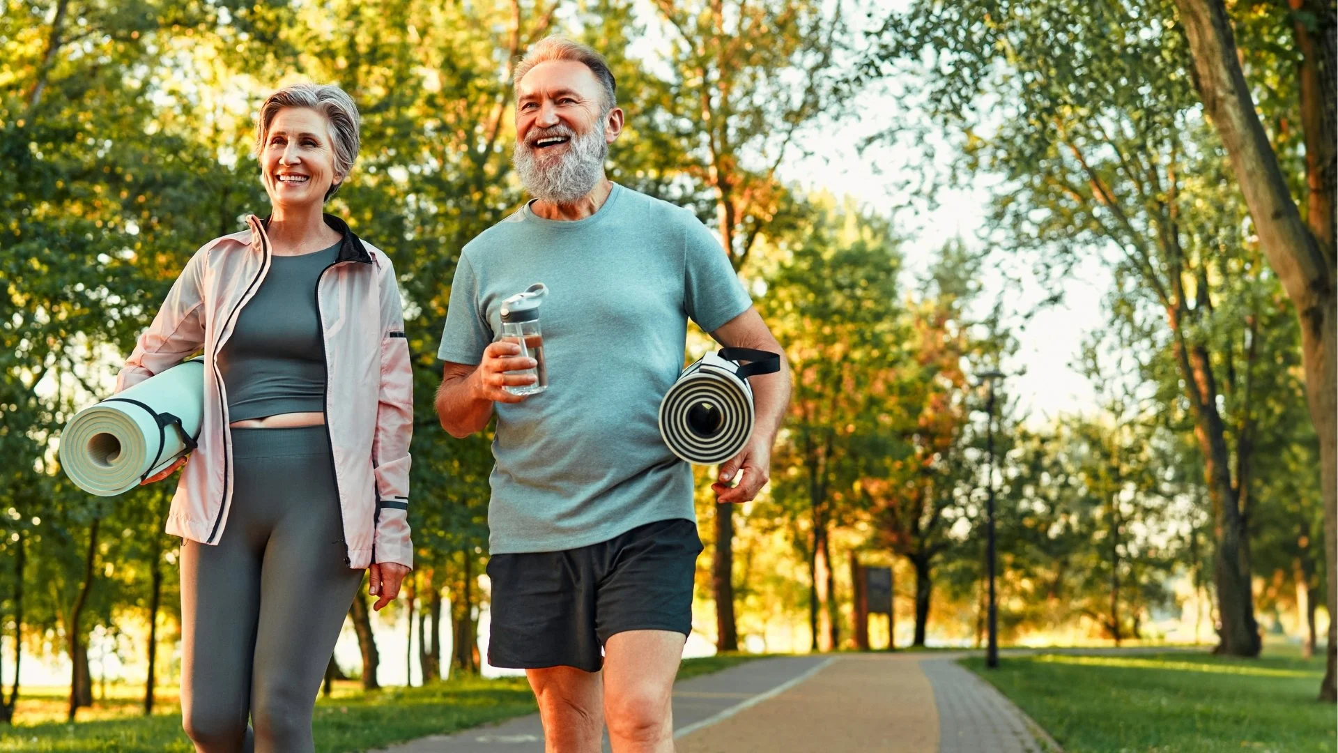 An older man and woman walking in a park with fall foliage, carrying yoga mats and water bottles, smiling and enjoying the outdoors.