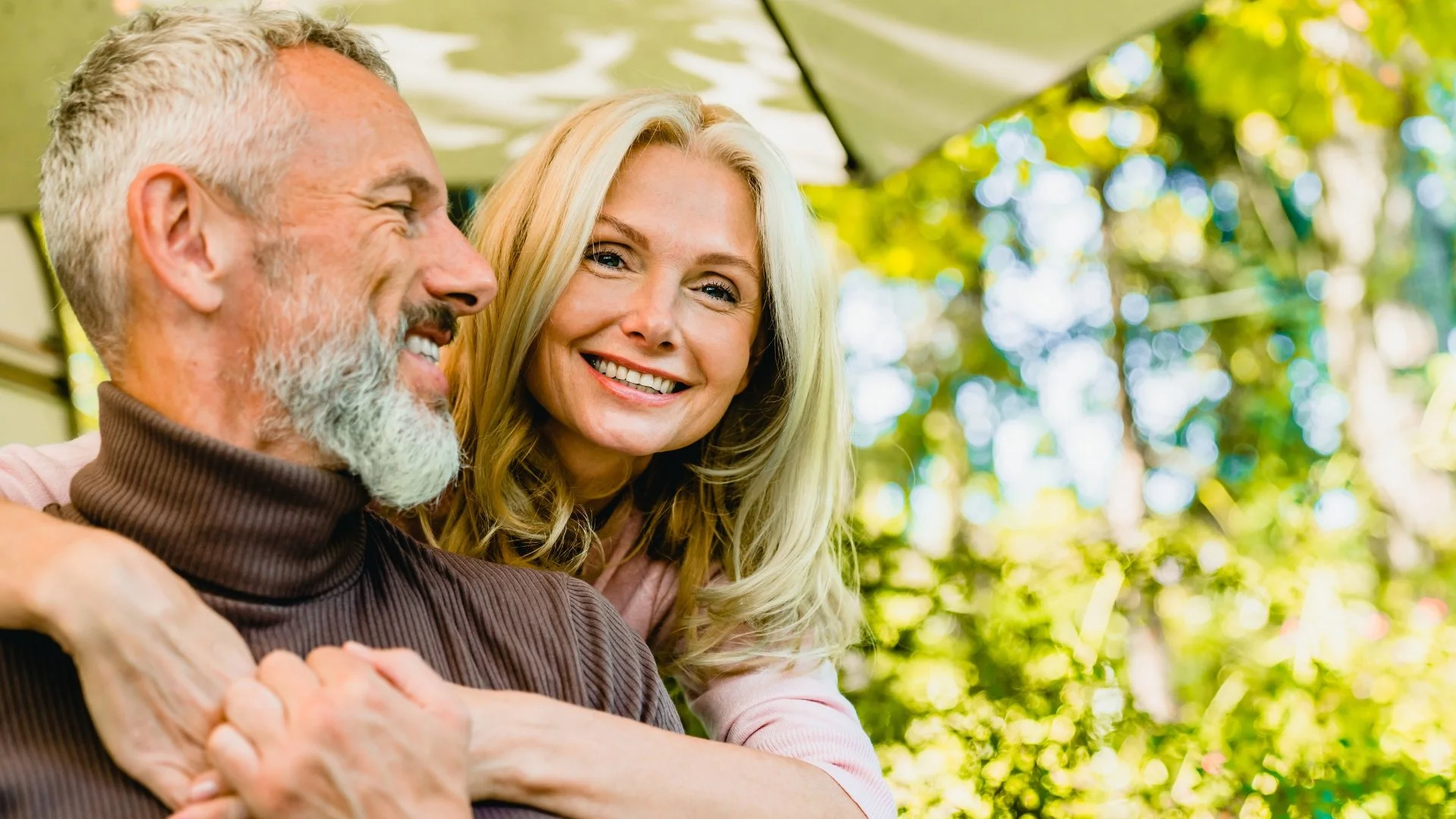 A smiling woman with blonde hair and a muscular older man with a beard sitting outdoors in a park or garden area, with trees and sunlight in the background.