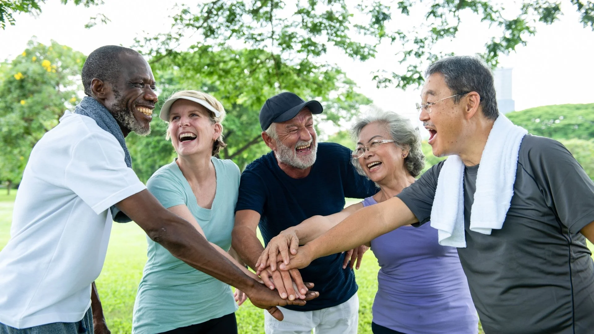 A group of diverse elderly people and a younger man in sports attire are outdoors, smiling and laughing, with their hands stacked together in a show of team spirit.