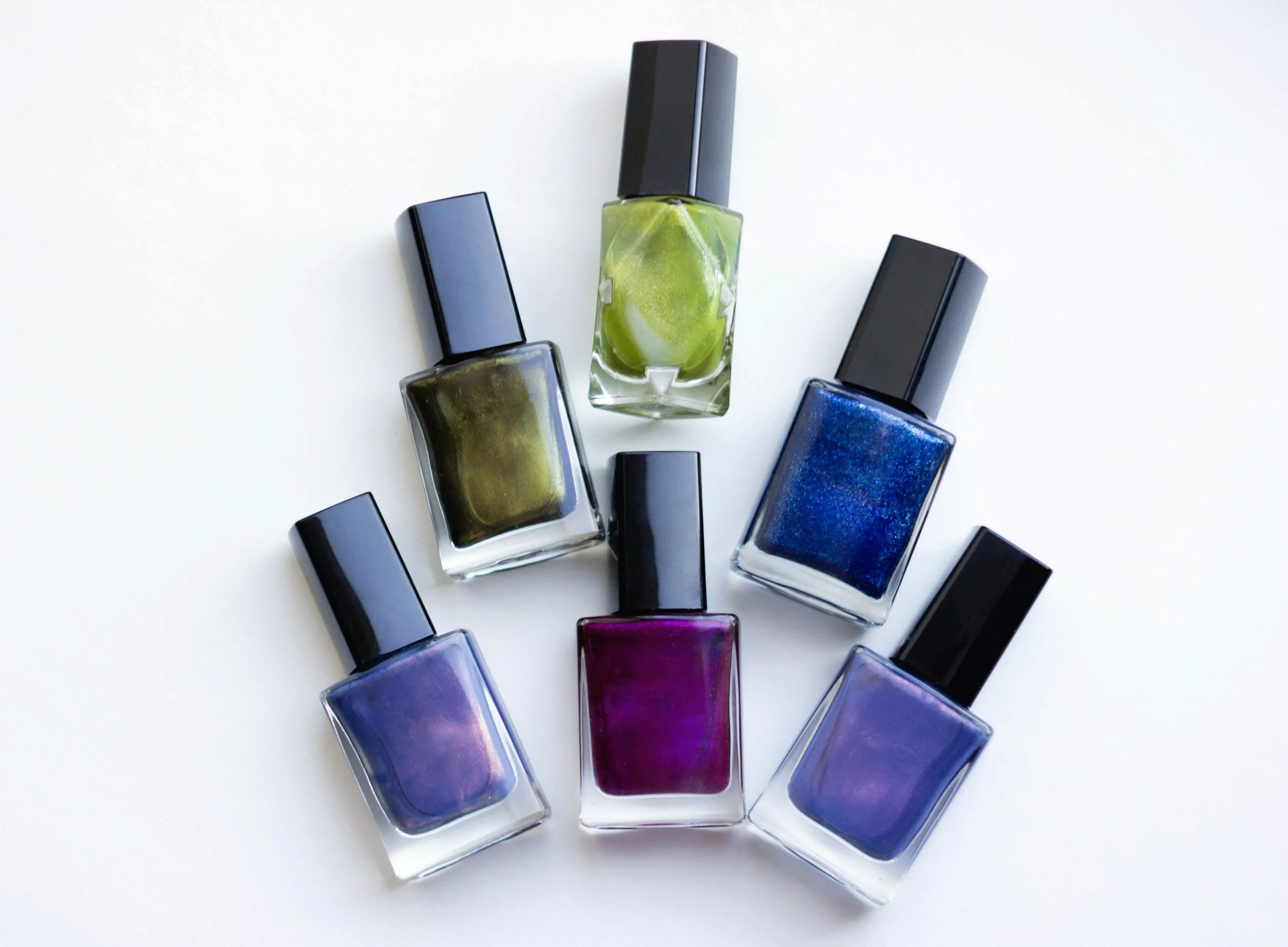 A collection of twelve bottles of nail polish arranged in a semi-circle on a white surface, featuring colors such as purple, blue, green, yellow, and red.