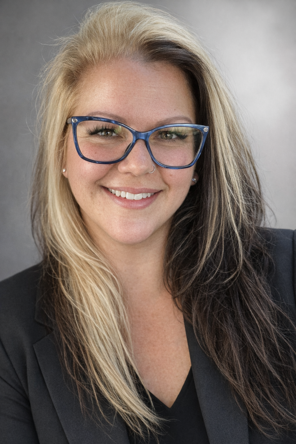 Close-up portrait of a smiling woman with blonde and dark brown hair, wearing blue glasses, earrings, and a black blazer with a dark top, against a neutral gray background.