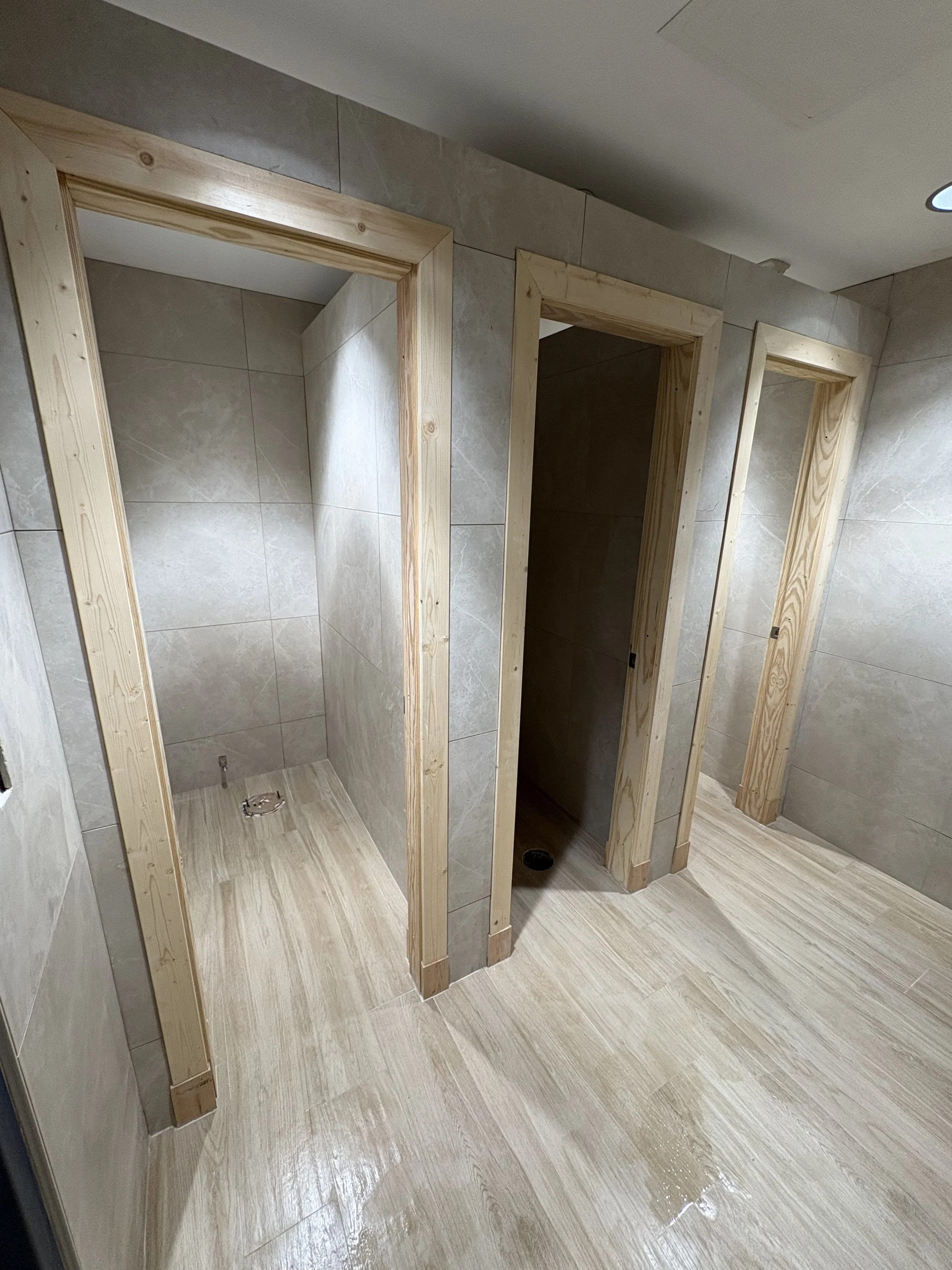 Construction site of three bathroom stalls with incomplete door frames, tiled walls, and a wet floor.