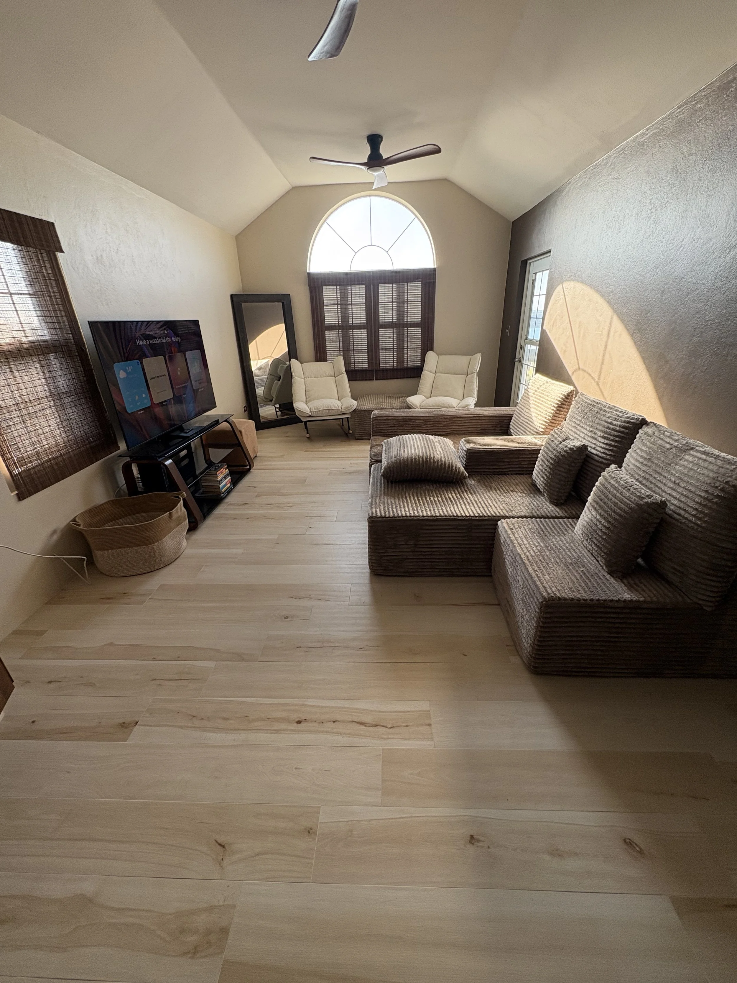 Living room with beige walls, light wood flooring, a large window with brown shutters, two beige armchairs, a brown sofa with matching cushions, a flat-screen TV on a stand, and a full-length mirror.