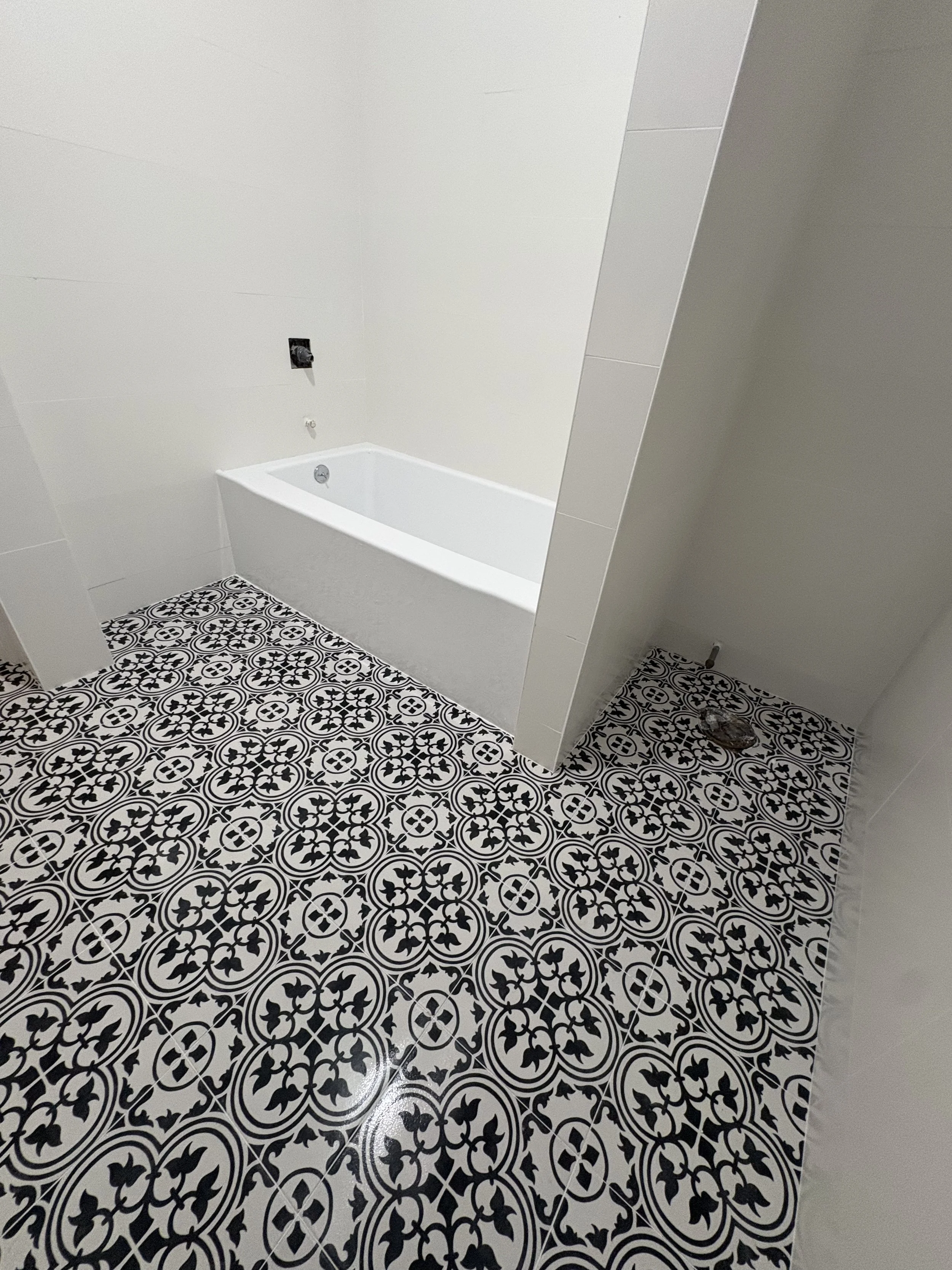 Bathroom with black and white decorative patterned floor tiles, white walls, a white bathtub, and a black wall-mounted fixture for a shower or faucet.