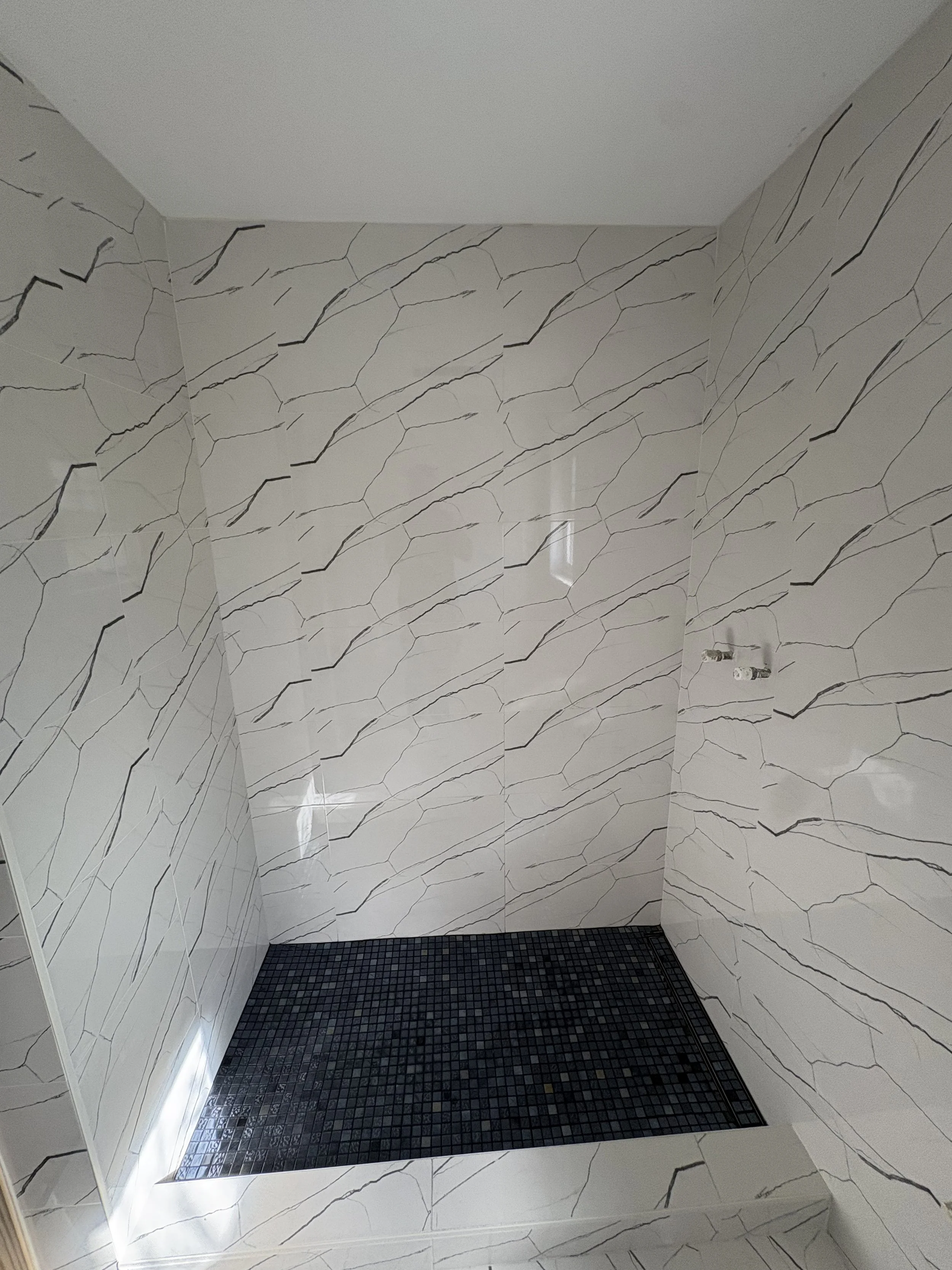 Bathroom shower with black mosaic tile floor and white walls with black veining, partially visible mirror on the left, and plumbing fixtures on the right wall.