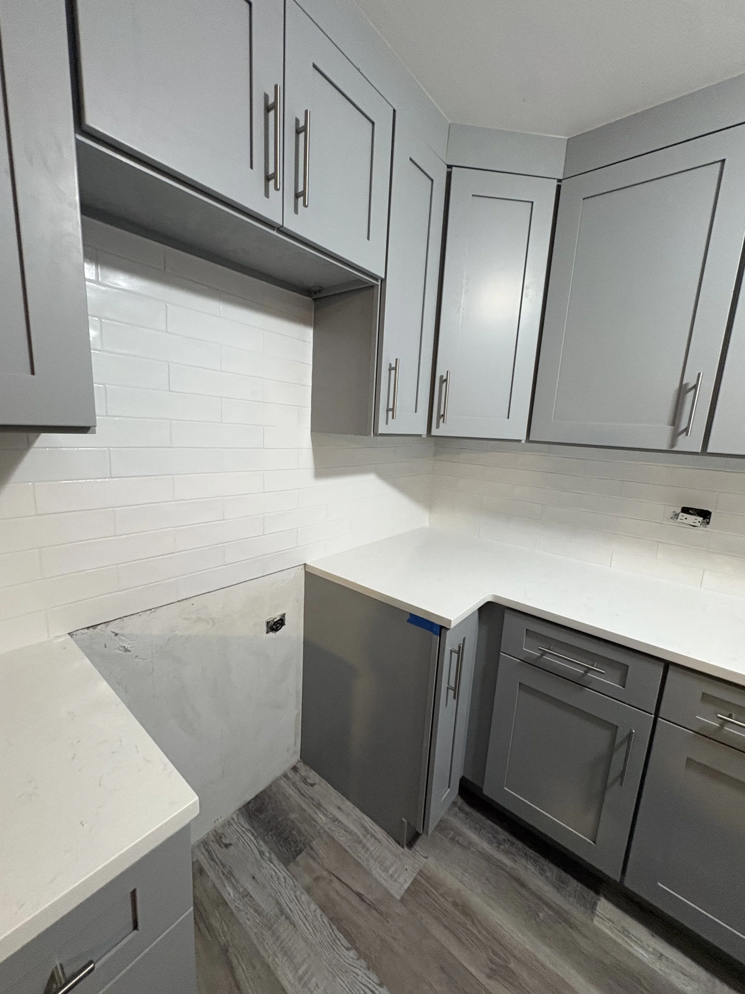 Kitchen with gray cabinets and white countertop, white subway tile backsplash, electrical outlets, and wood-look vinyl flooring.