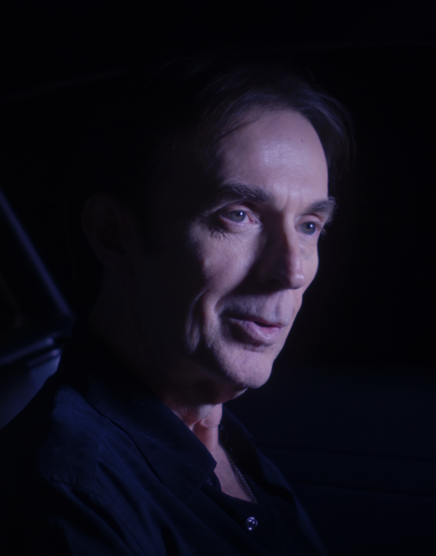 Close-up of a man with dark hair and blue eyes, looking to the side, in a dark environment with subtle blue lighting.
