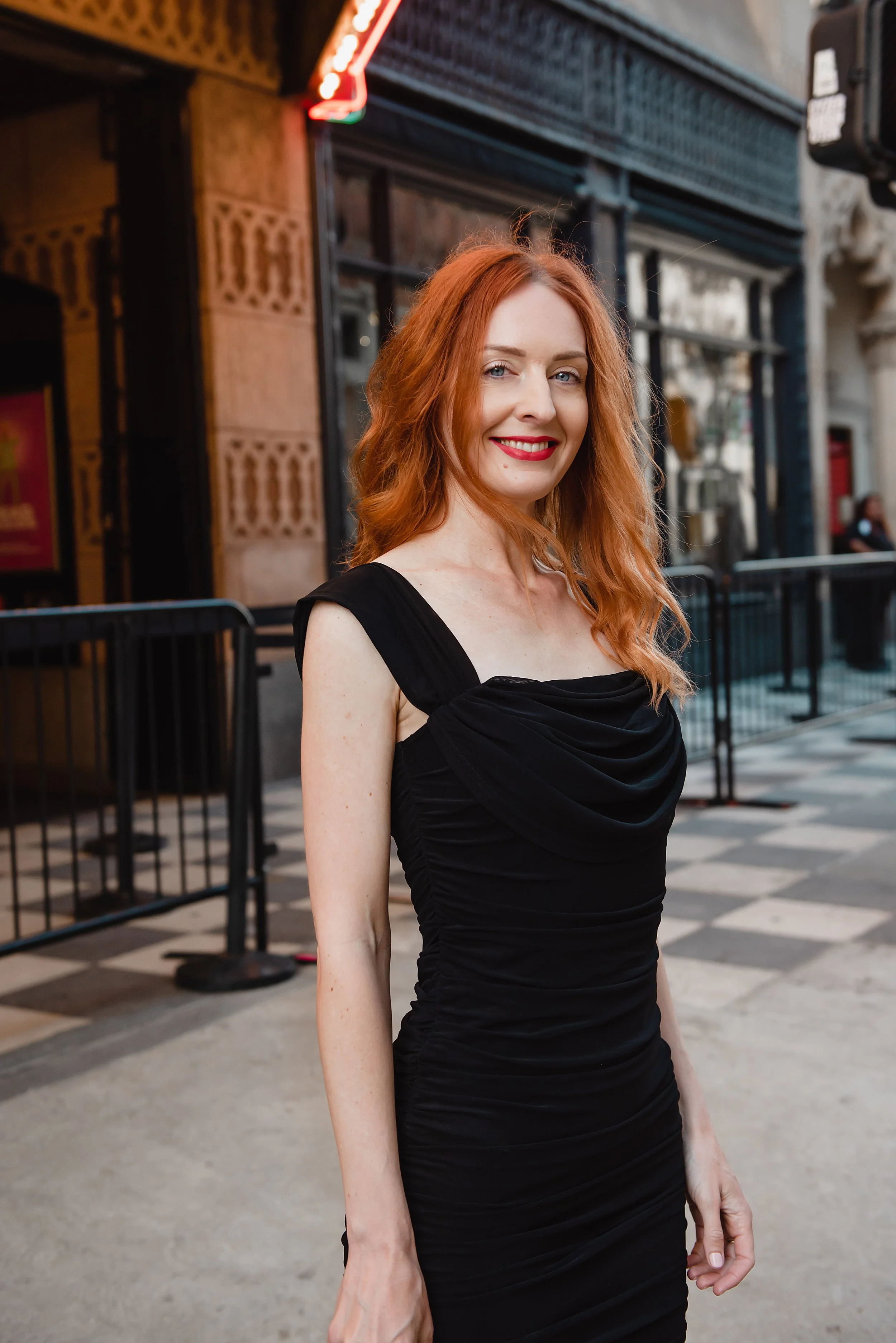 A smiling woman with red hair wearing a black dress standing on a city street with a building and a person with a mask in the background.
