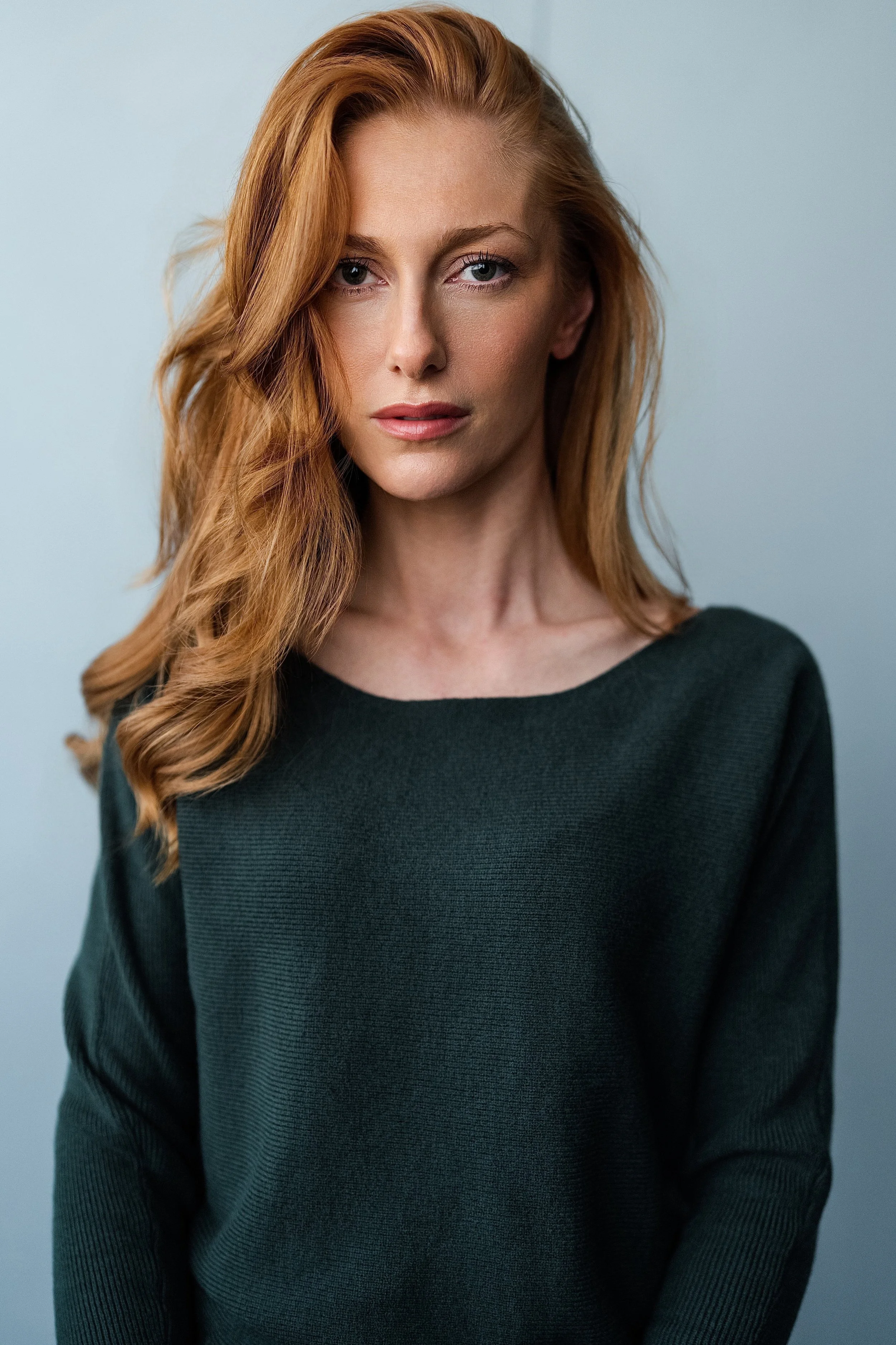 A woman with long, wavy red hair and fair skin wearing a dark green top, standing against a plain light blue background.