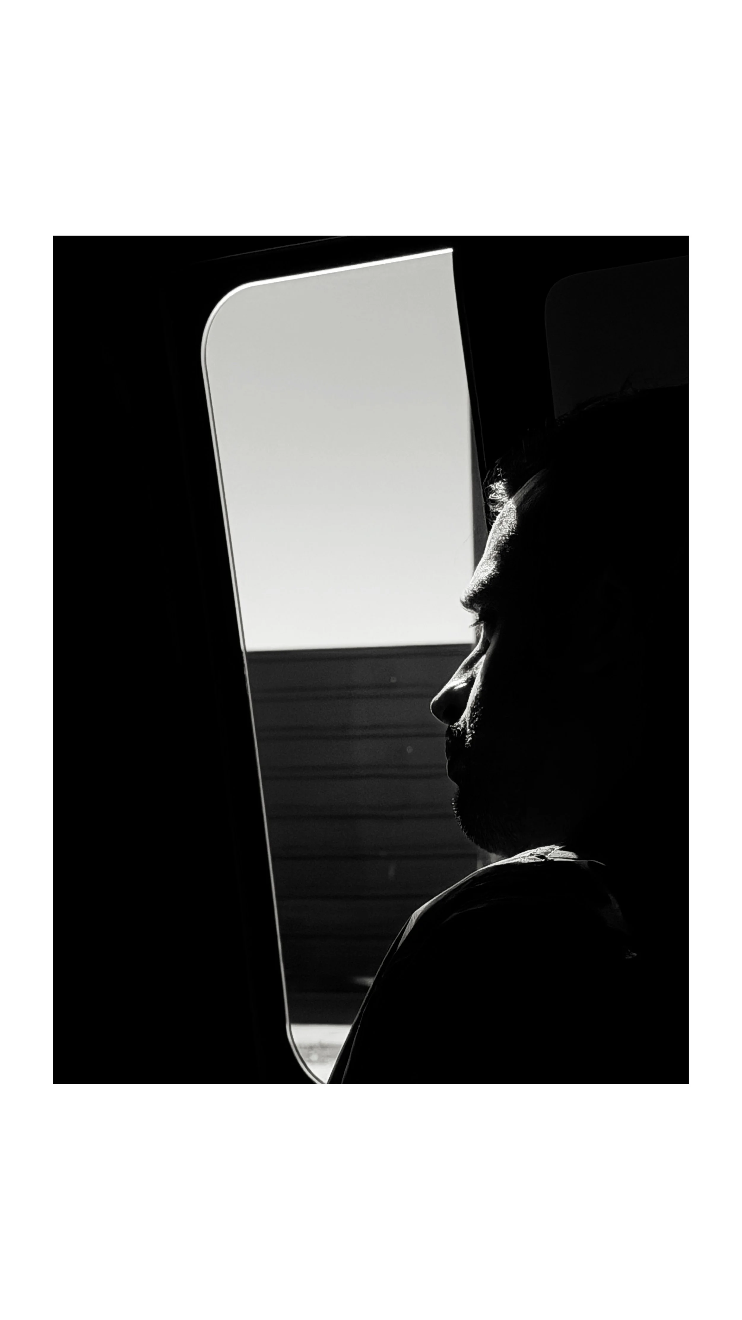 Black and white photo of a man with dark hair and beard, sitting in front of a computer screen, illuminated by the light from the monitor. The background has horizontal lines suggesting a window or panel.
