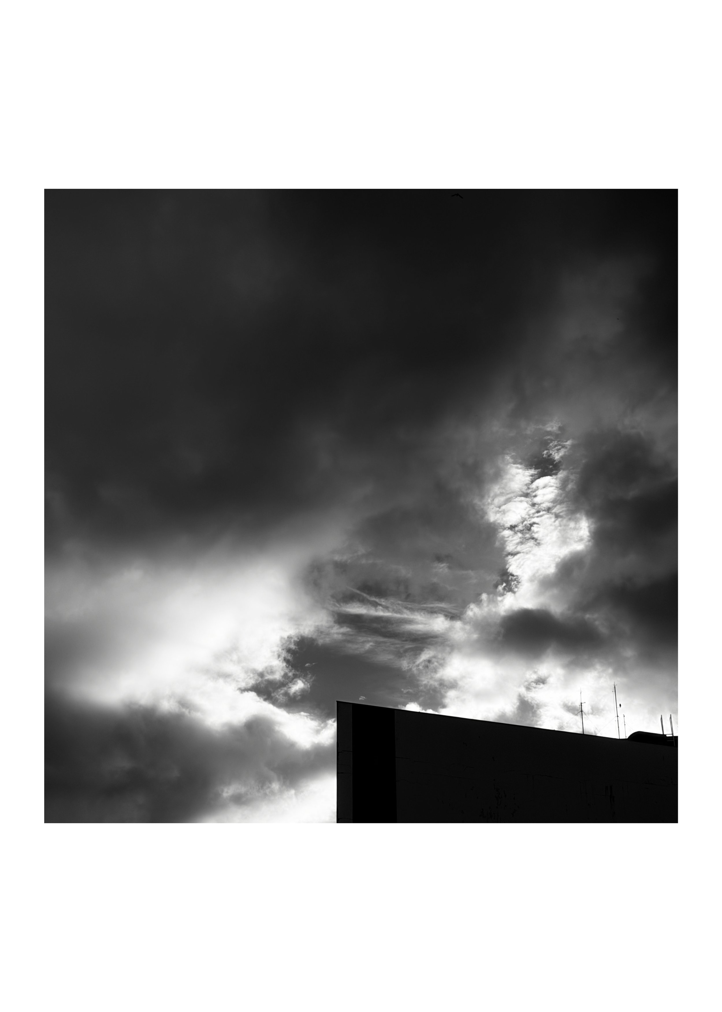 Dark, cloudy sky with patches of sunlight breaking through clouds, part of a building with antennas at the bottom right.