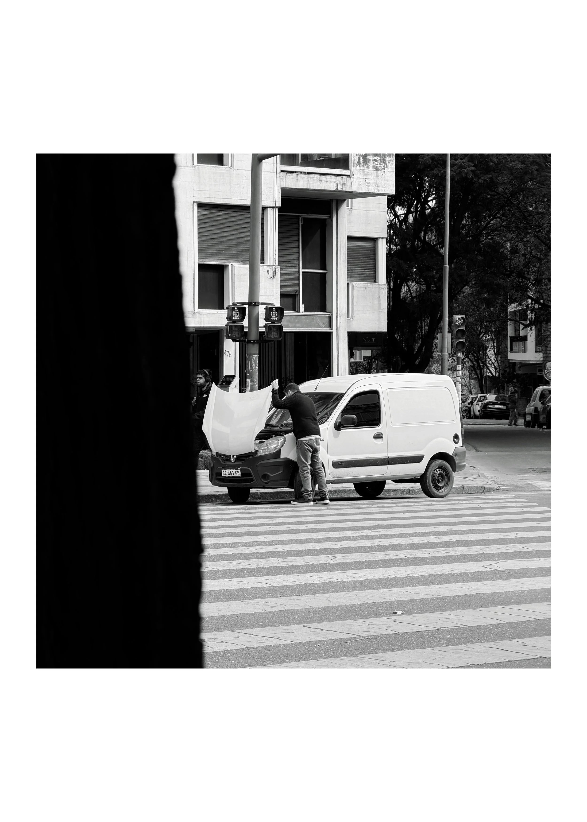 A black and white street scene with a person working on a white van at a crosswalk, buildings, traffic lights, and trees in the background.