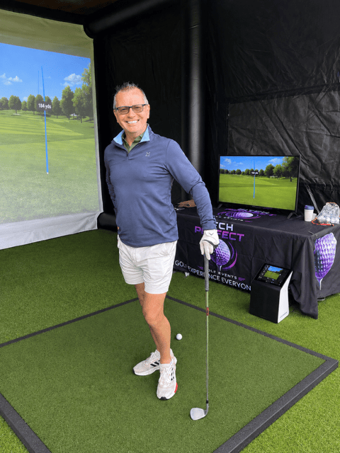 A male adult stood inside an inflatable golf bay leaning on a golf club. Also showing an image of a golf course on the impact screen at the back of the bay and on a TV screen at the side of the bay