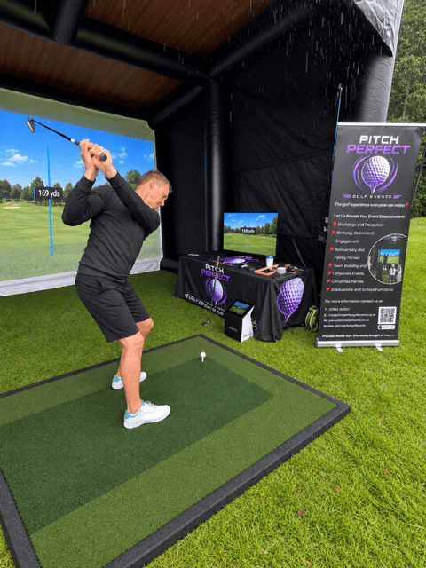 A male adult swinging a golf club inside an inflatable golf bay. Also showing an image of a golf course on the impact screen at the back of the bay and on a TV screen at the side of the bay