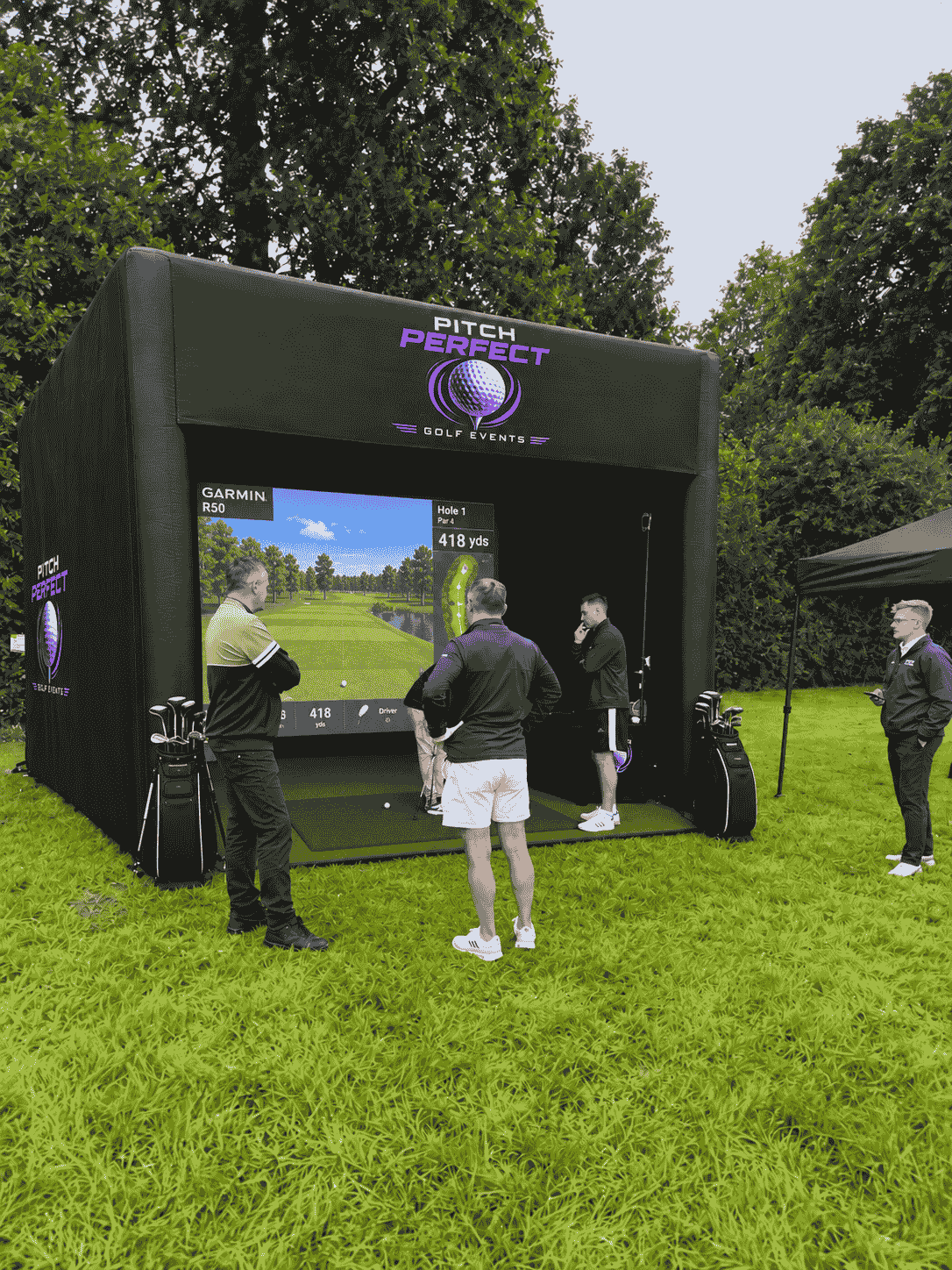 A group of people standing in and around an inflatable golf bay awaiting their turn to hit a golf ball. At the back of the golf bay is a screen showing an image of a golf hole
