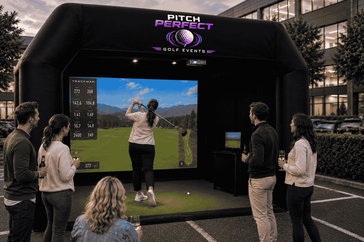 Office colleagues in a car park  watching a team member swing a golf club within an inflatable golf bay. There is a screen that shows how far she has hit the ball and where the ball has landed